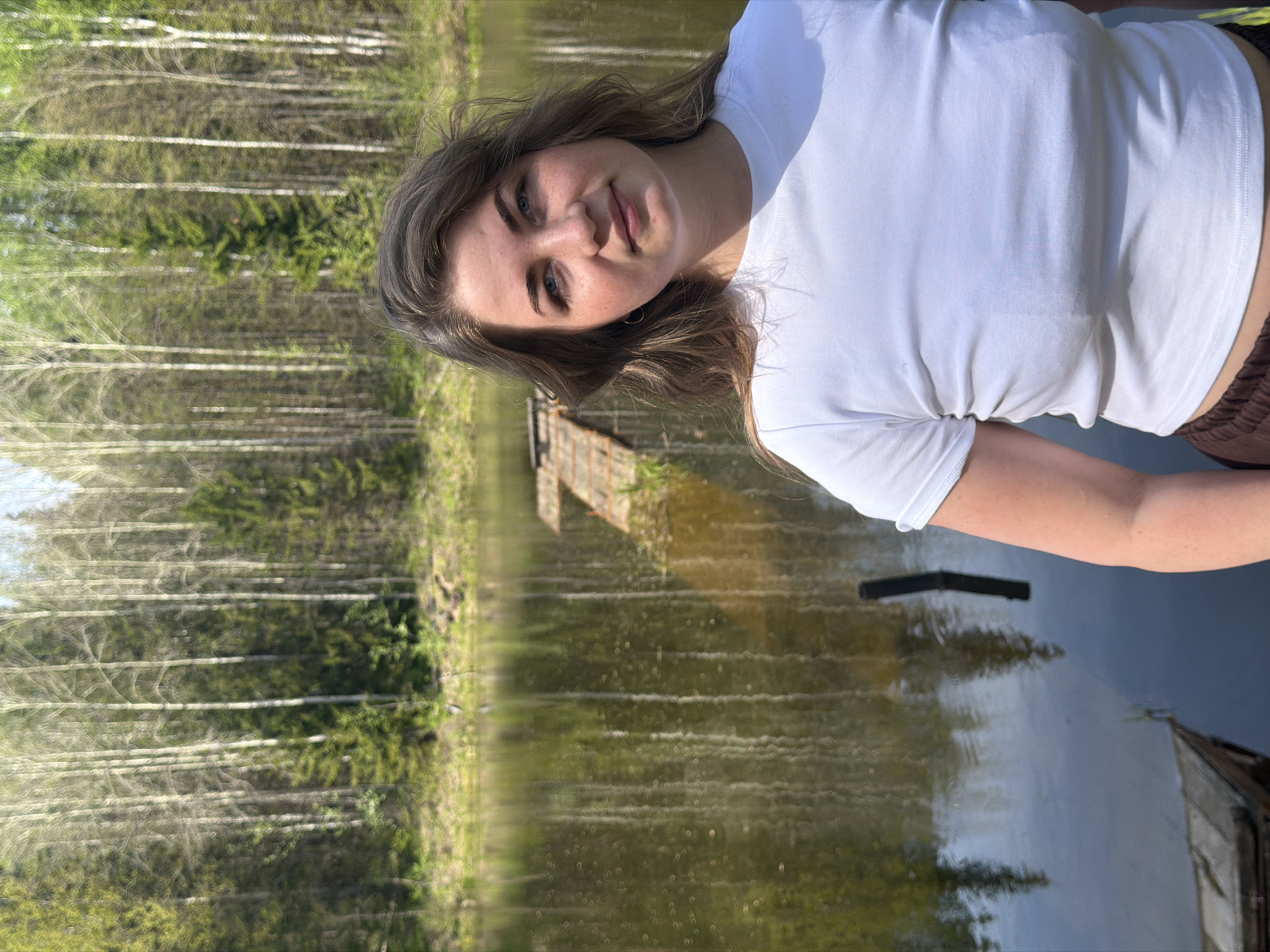 A woman with wavy brown hair and wearing a white t-shirt poses for a photograph outdoors next to a body of water. The background includes trees and reflections in the water. A wooden structure is visible on the water. The photograph was taken in Ural'skoe, Russia.