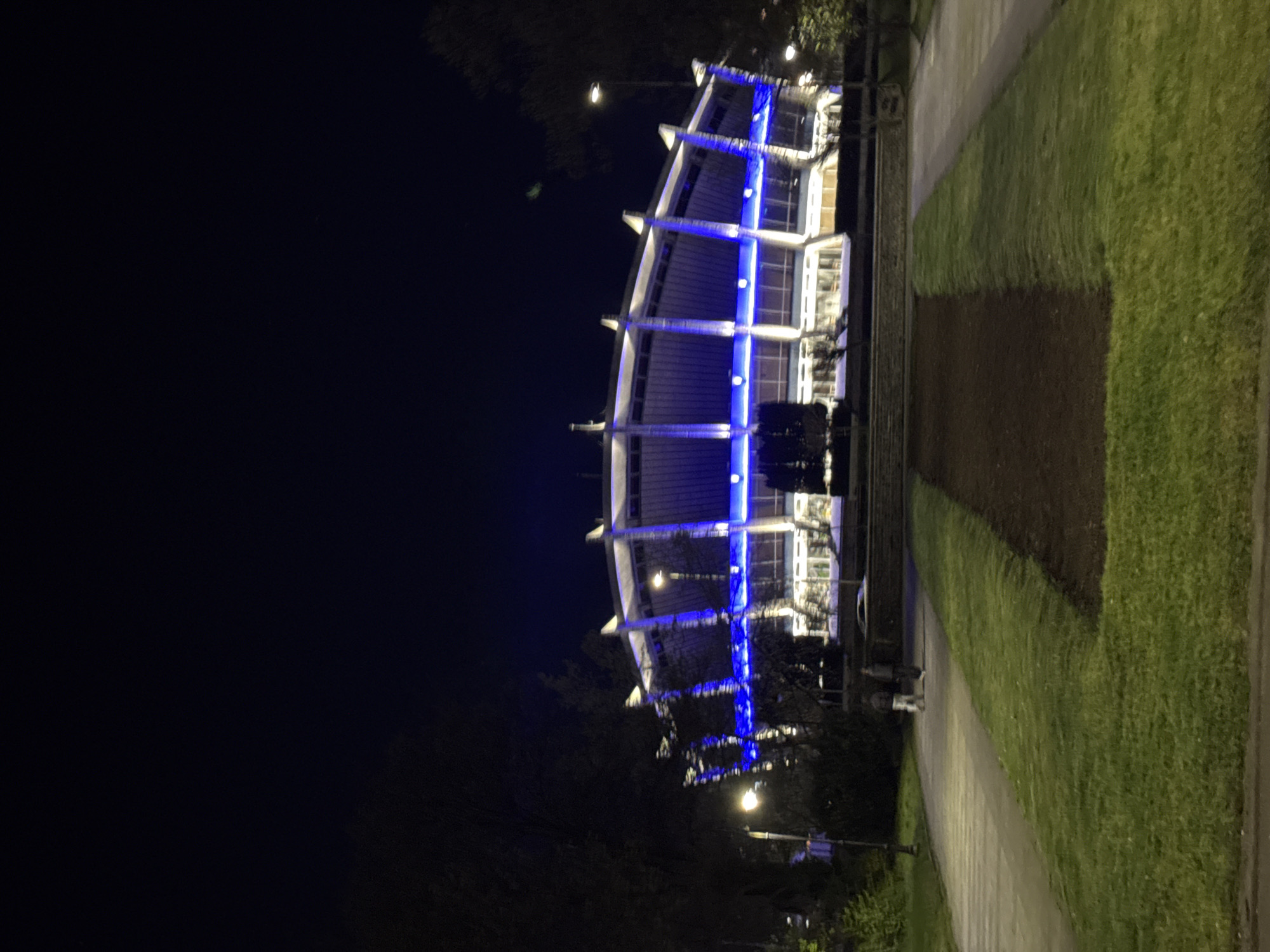 The image depicts a large, distinctive building illuminated with bright blue lights against a dark night sky. The multi-story structure features a curved, modern facade with prominent white architectural supports and large windows. Horizontal strips of vibrant blue LED lighting accentuate the building's form, running along its exterior.
In the foreground, a paved pathway extends horizontally, flanked by areas of green grass. A strip of darker ground, possibly a garden bed or recently turned soil, lies between two sections of manicured lawn. Several streetlights are visible, casting warm, yellow-toned light onto the path and surrounding greenery, creating a contrast with the cool blue illumination of the building.
Dark trees and foliage are present in the background and along the edges of the scene, indicating a landscaped setting. The time of day is clearly night, with the sky appearing completely dark. No specific activities are taking place, and no people or visible text are discernible in the image. The architectural style and prominent lighting suggest a public or institutional building, such as a cultural center or sports venue, in Varna, Bulgaria.