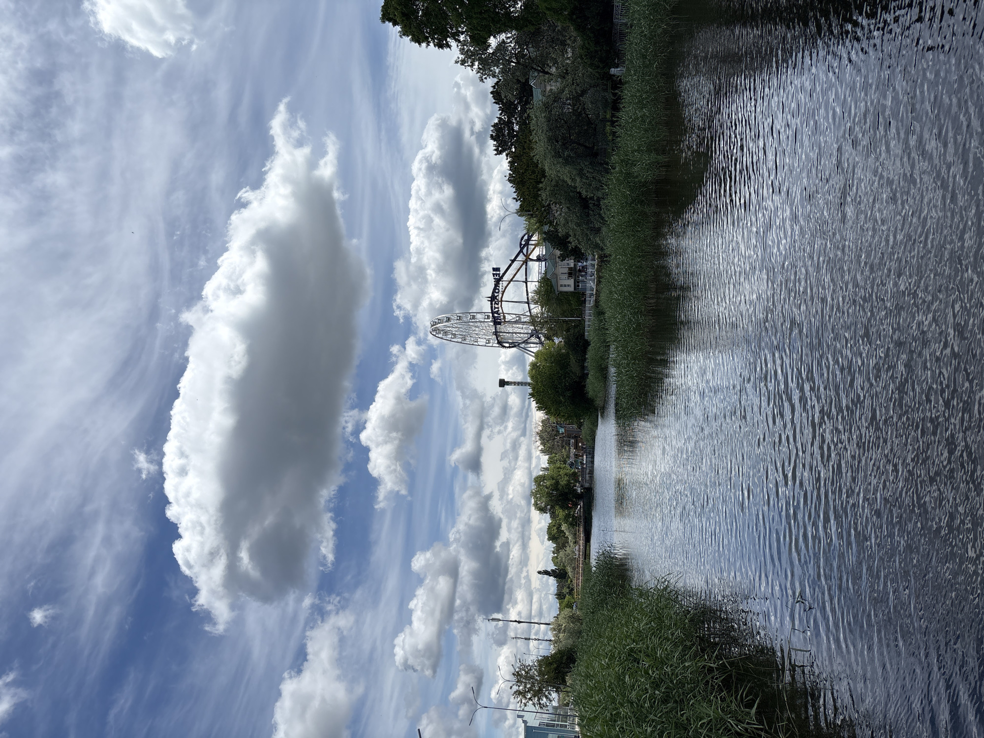 The image depicts a canal, potentially the Anapka River, in Anapa, Russia, flanked by dense reeds and trees. Visible in the background are elements of an amusement park, including a Ferris wheel and a roller coaster. The sky above is filled with cumulus clouds against a blue backdrop. The water in the canal is rippled, reflecting light and shadows. No people are apparent in the image.