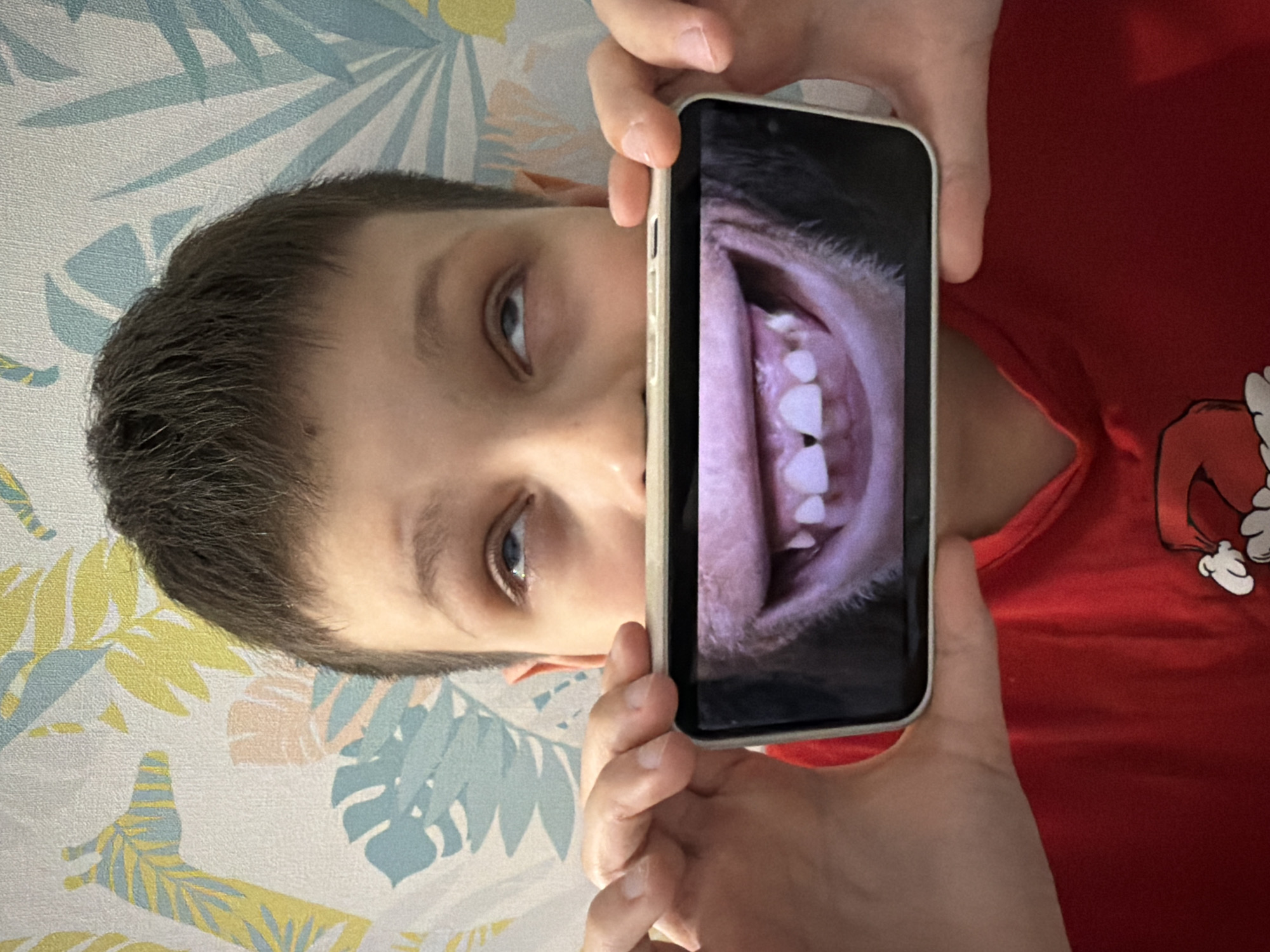 A young boy is depicted in a sideways orientation, appearing to be lying on a patterned surface while holding a smartphone. He has light skin, short dark hair, and blue eyes, and is looking towards the phone. He wears a red t-shirt, part of which is visible, featuring a white cartoon-like design near the right shoulder area.
The boy holds a smartphone, encased in a light-colored cover, with both hands in front of his face. The phone's screen displays an extreme close-up, somewhat distorted image of an open mouth, showcasing white teeth, pink gums, and the interior of the mouth. The detail on the phone screen is very pronounced, focusing intently on the oral cavity.
The background is a light-colored surface, possibly bedding or wallpaper, adorned with a repeating pattern of stylized leaves in shades of light blue, yellow, and soft peach. This suggests an indoor setting, likely a room. The activity centers around the boy's interaction with the phone, potentially taking an unusual selfie of his mouth, examining dental features, or viewing a macro image. No specific time of day or weather conditions are evident. The location for this scene is Votkinsk, Russia. There is no visible text in the image.