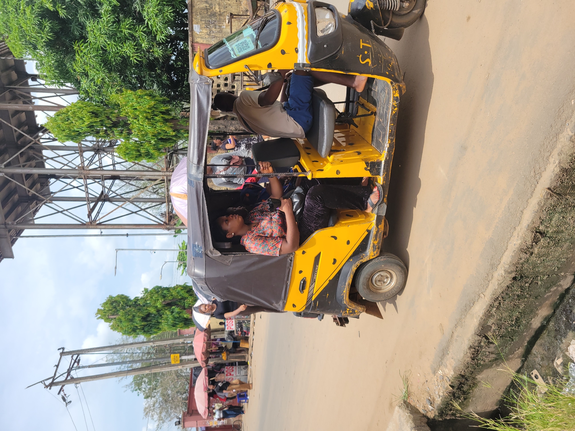 The image shows a yellow and black auto rickshaw (a three-wheeled vehicle) in Egbagu, Nigeria. The auto rickshaw is carrying passengers, including a woman in a patterned shirt and black pants, sitting in the back. The driver, a dark-skinned man, is in the front seat. The rickshaw is on a paved road, with pedestrians and stalls visible on one side. A metal structure looms overhead. The day appears sunny. A drainage ditch runs along the road's edge.