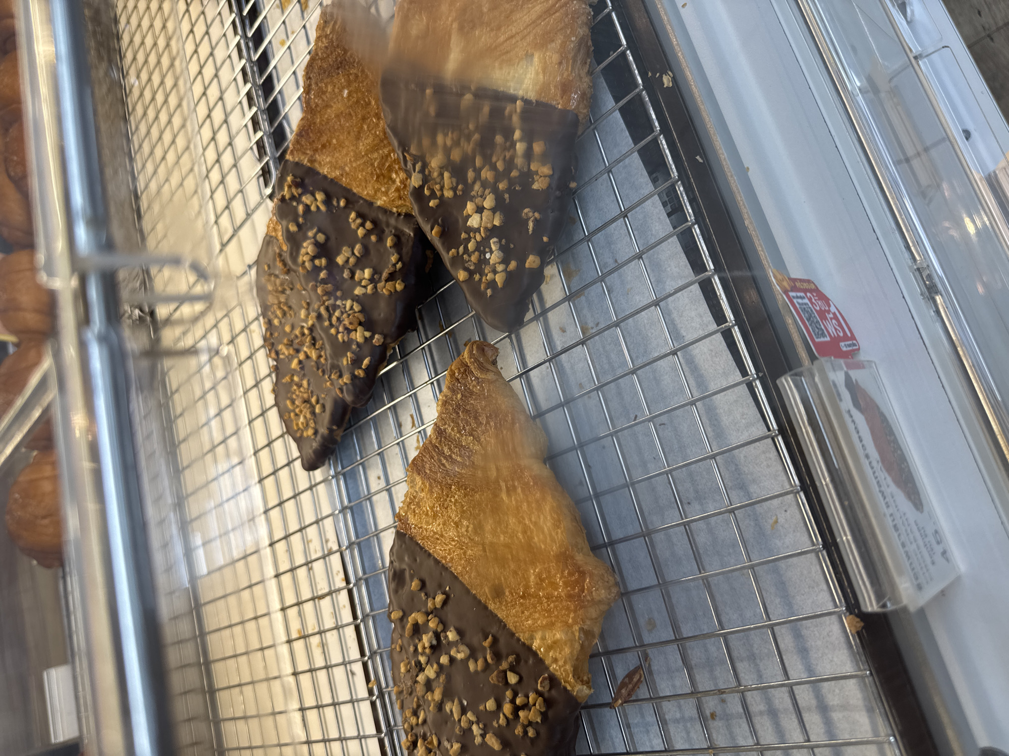 This image shows a close-up view of pastries in a refrigerated display case, likely in a bakery or cafe in Bangkok, Thailand. The pastries are croissants that have been dipped in chocolate on one half and sprinkled with chopped nuts. They are arranged on a metal cooling rack. In the background, other baked goods, possibly more croissants, are visible. A price tag or menu item label is partially visible in the lower left corner, showing the price "45" in Thai Baht. The lighting suggests an indoor setting, possibly a shop or market.