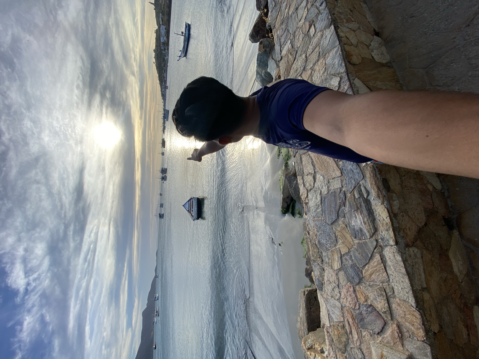 The image shows a man pointing towards the ocean in Juan Griego, Venezuela. The man has dark hair and is wearing a blue shirt. He is standing on a stone structure. In the background, there is water with several boats, and the sky is partly cloudy. The sun is shining brightly.
