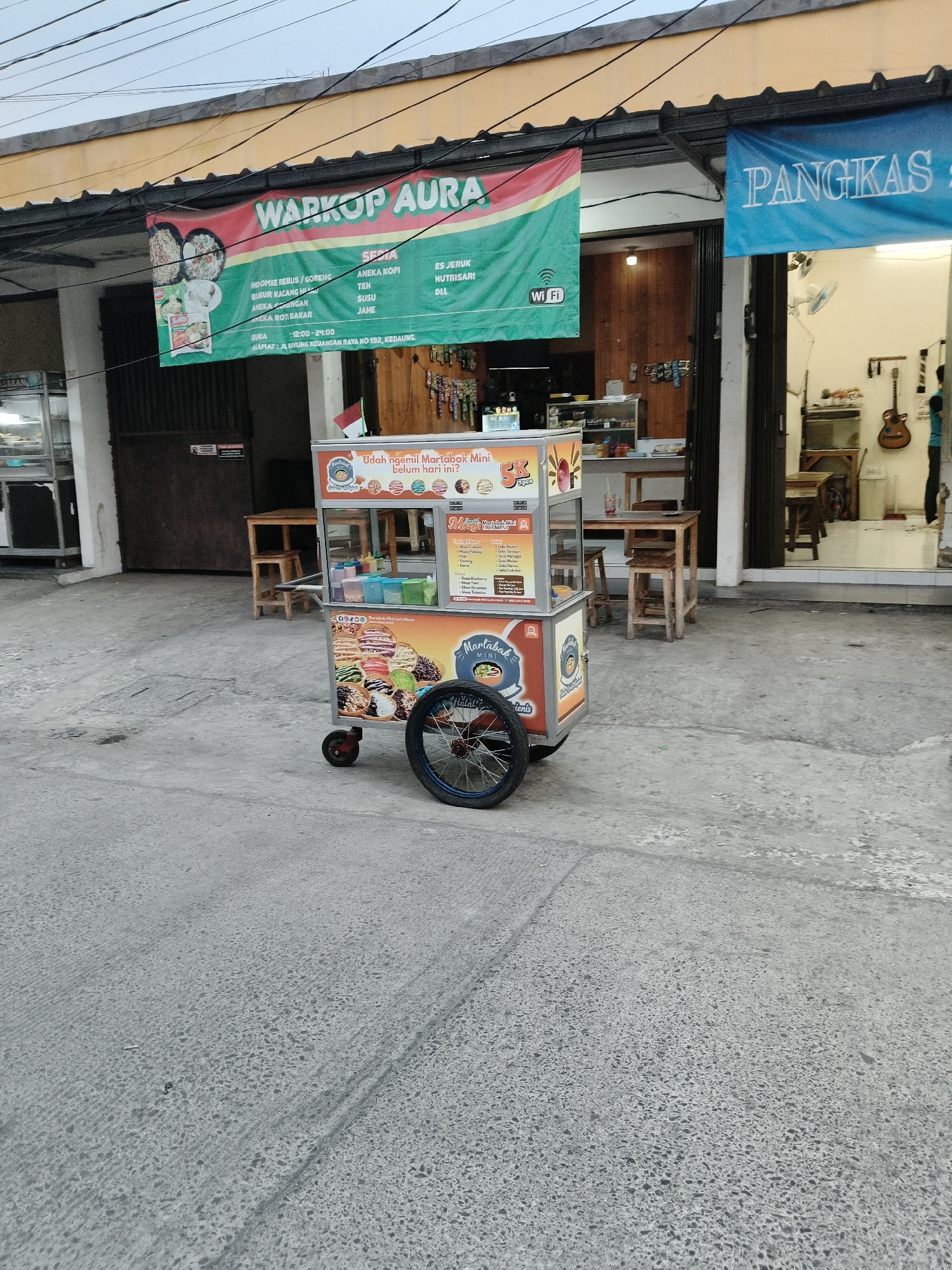 The image depicts a street scene in South Tangerang, Indonesia, with a martabak mini cart as the focal point. The cart, a mobile food stall, is positioned on a paved road, its colorful signage advertising various martabak flavors. Behind the cart, a building with shops is visible, featuring banners for a "WARKOP AURA" and "PANGKAS". The area appears to be an urban commercial space, with tables and chairs set up outside the shops, suggesting a place for patrons to dine or relax. The sky above is overcast, indicating a cloudy day. No people are prominently featured in the scene, with only a partial view of a person standing inside one of the shops. The overall impression is that of a typical street-side food vendor operation in a residential or commercial district of South Tangerang.