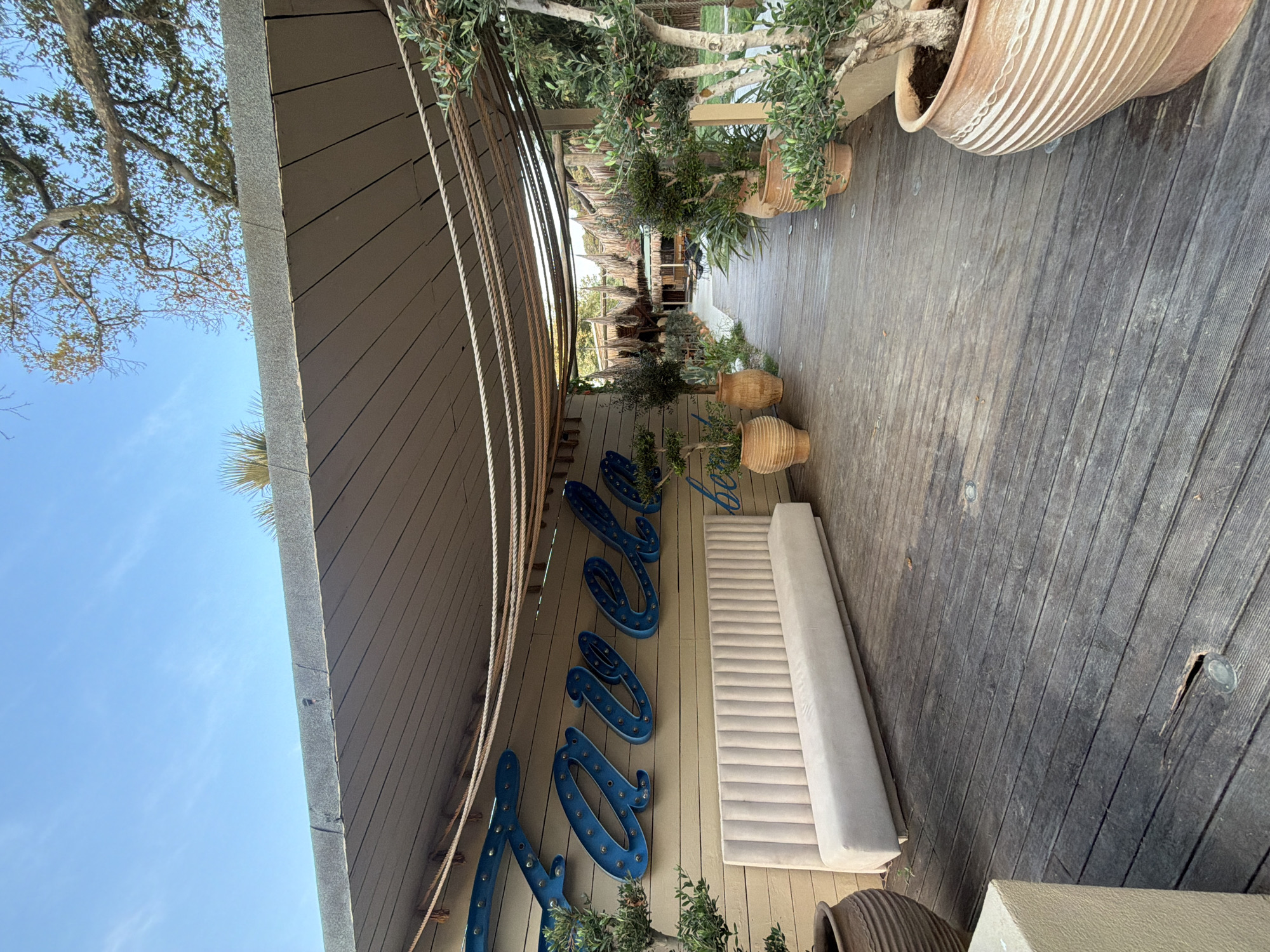 A wooden walkway leads through an outdoor area with a thatched roof structure visible in the distance. On the left side of the walkway, a light-colored tufted bench is placed against a wooden wall adorned with blue neon letters that spell out "ZAFELO BEACH". Several potted plants, including olive trees, are situated along the walkway. The sky is clear and blue. The scene appears to be a tranquil outdoor seating or dining area.