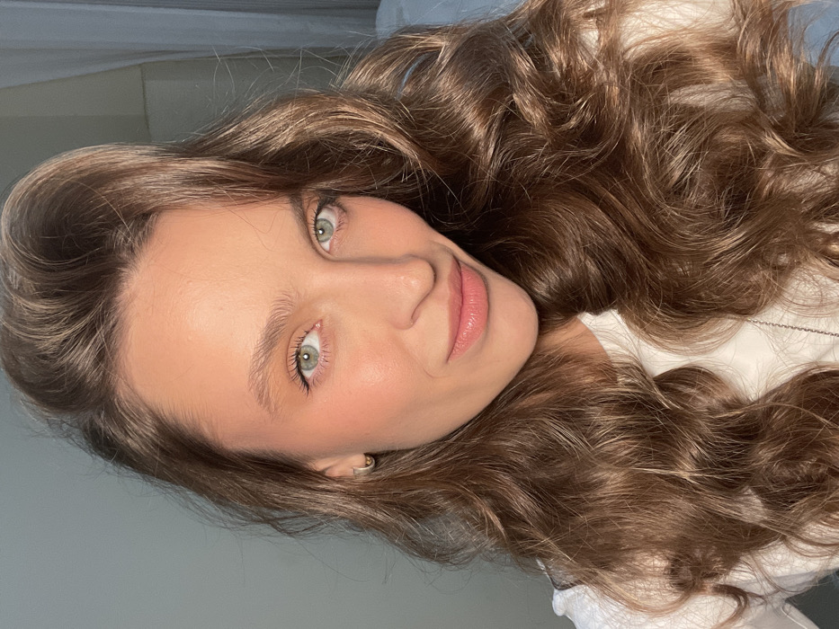This close-up portrait features a young woman with long, voluminous, wavy brown hair spread around her. She has striking light green or blue-green eyes that look directly at the viewer, and a soft, natural expression with a hint of a smile. Her skin appears smooth, with a subtle blush on her cheeks and a natural pink tint on her lips. She is wearing a white top, possibly a shirt or blouse, and a thin silver chain necklace is visible near her collarbone. The background is soft and out of focus, suggesting an indoor setting with diffused lighting. The overall mood is serene and natural. The image is taken in Budva, Montenegro. There is no visible text.