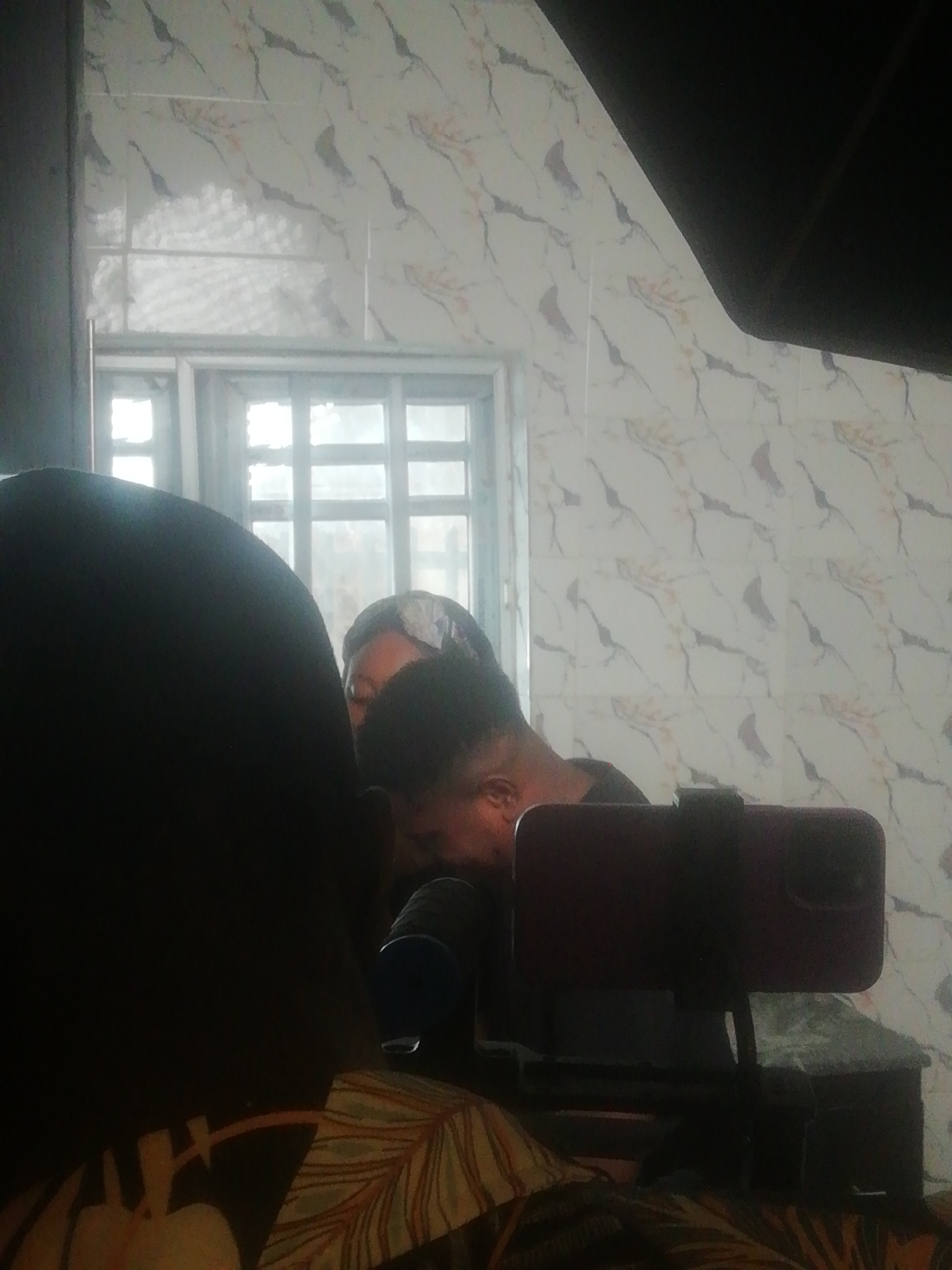 The image depicts an indoor scene in Ikorodu, Nigeria, where at least two individuals appear to be engaged in recording or live-streaming content. In the foreground, the back of a person's head and shoulder are largely silhouetted, dressed in clothing featuring a green, orange, and white leaf pattern. To their right and slightly behind them, another person is visible, with their head tilted downwards, showing part of their ear and side profile. A third person's forehead and eyes are vaguely discernible behind the foreground individual's head.
A dark-cased smartphone, likely recording, is mounted horizontally on a stand in the middle-right of the frame. Adjacent to the phone stand, a blue and silver/grey microphone is partially visible, indicating audio capture. The background features a tiled wall adorned with a light-colored, marble-like pattern comprising white/cream tones with dark grey/black and orange/brown streaks. A multi-paned window, letting in bright daylight, is situated behind the subjects, suggesting the activity is taking place during the day. A dark, possibly overhead, structure is visible in the upper right corner of the frame. No discernible text is visible in the image.