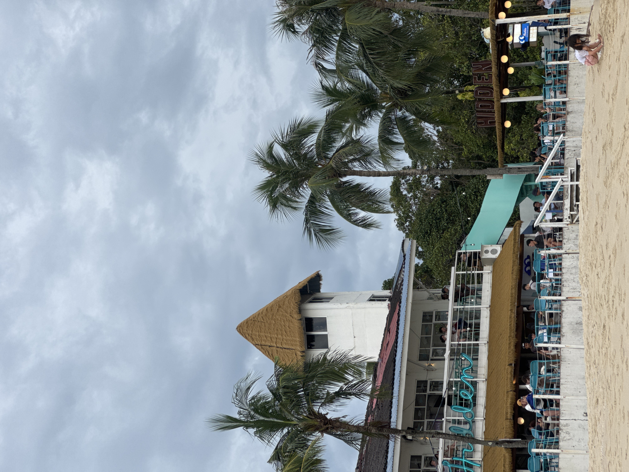 The image depicts a beachfront establishment on Tengah Island, Malaysia, characterized by a two-story building with a distinctive thatched conical roof on a white tower. Palm trees are prominent in the foreground and surrounding the building. The lower level of the establishment features an outdoor seating area with numerous tables and turquoise chairs, occupied by patrons. A neon sign spelling "Hidden" is visible on the upper level. Further along the beachfront, another section of the establishment displays a wooden sign reading "HIDDEN" above a covered seating area with glowing string lights. The scene is set against a backdrop of a cloudy sky and lush green foliage. In the foreground, a young girl is playing in the sand.