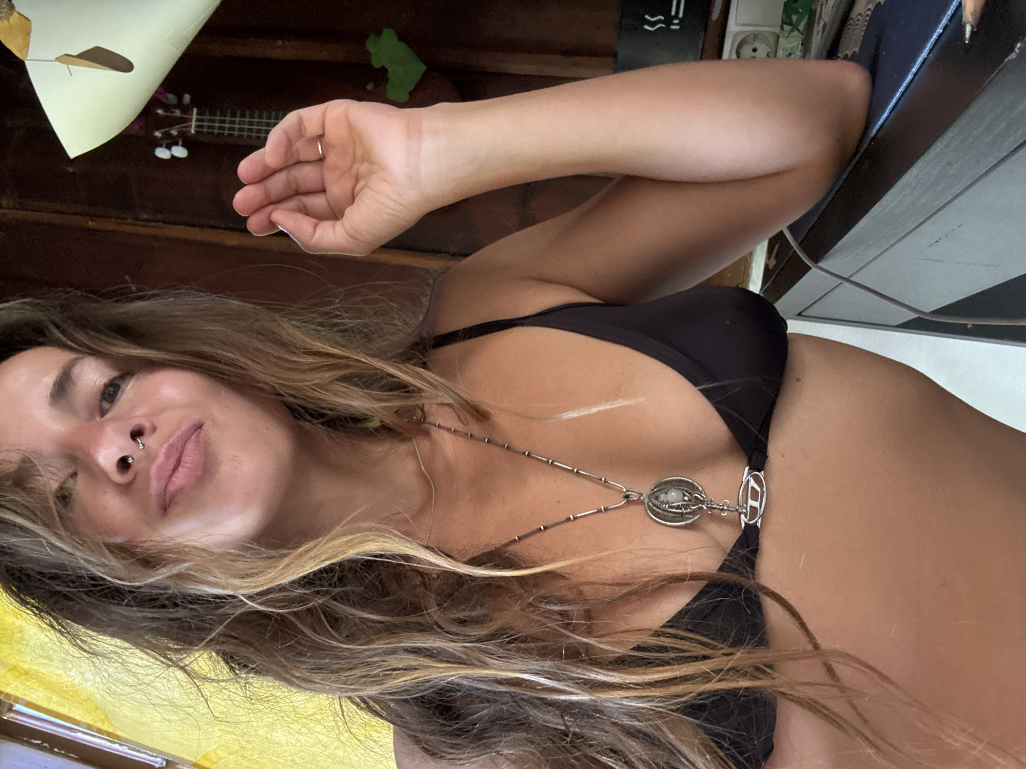 A woman is captured in a close-up, high-angle, rotoscope style shot. She has long, wavy, blonde-brown hair that frames her face, which is turned towards the camera with a slight smile. She has a septum piercing and her eyes are open. She is wearing a black bikini top with a silver necklace that has a unique pendant. The background is a dimly lit room with wooden paneling. On the wall behind her, a ukulele and a green object are visible. A yellow object is also present in the lower left corner of the frame. The lighting suggests it might be daytime, but the indoor setting makes it difficult to determine. The overall mood appears relaxed and casual.