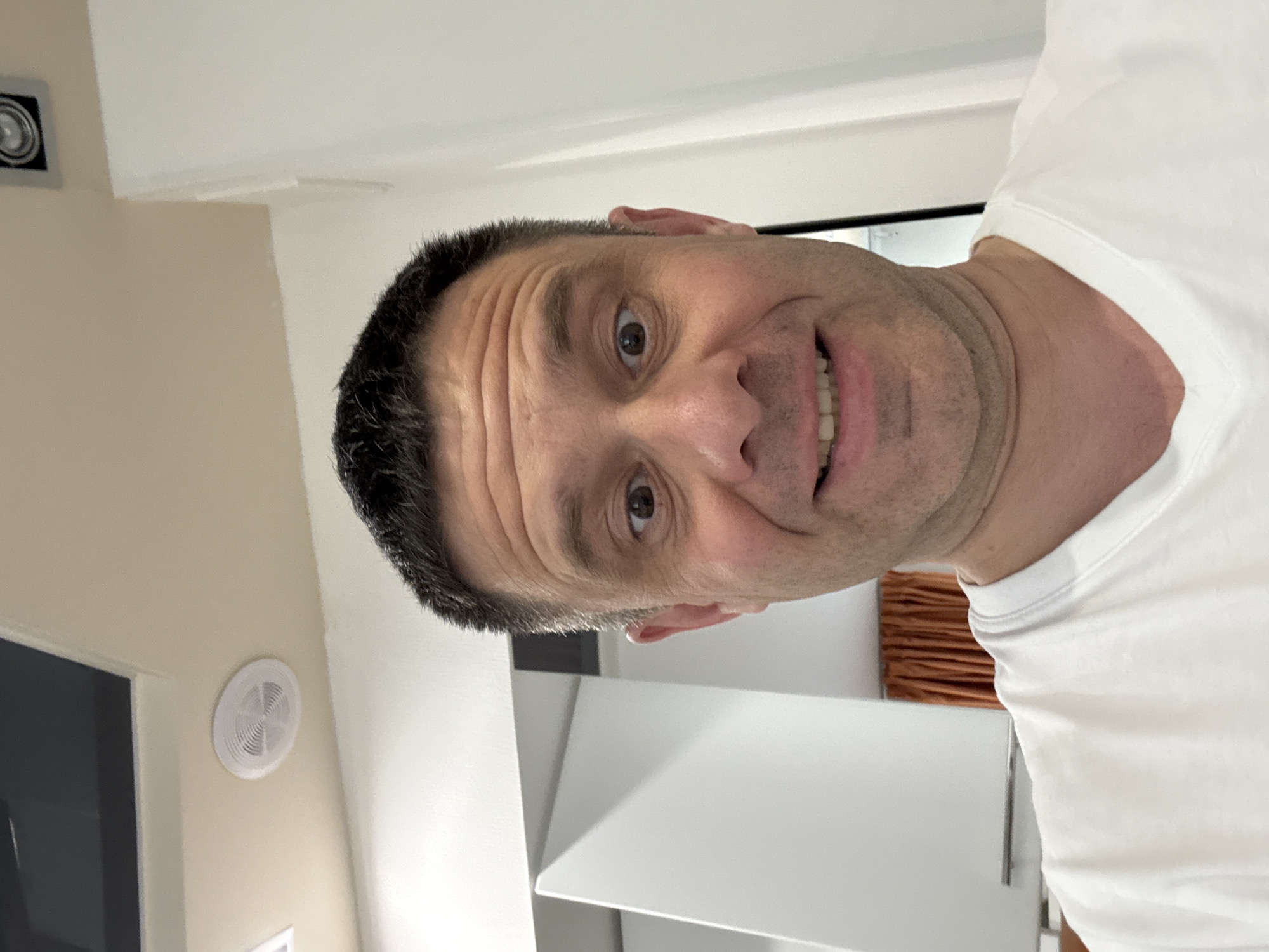 A close-up, slightly angled headshot of a middle-aged man with short, dark hair, light skin, and visible forehead wrinkles. He is wearing a white t-shirt. He is smiling slightly, exposing some teeth. The background shows a light-colored wall with a square light fixture near the top left corner and a round ventilation fan on the left side. A partially visible doorway and a white wall are also present. The location in the city of Turgoyak, Russia cannot be determined from the image.