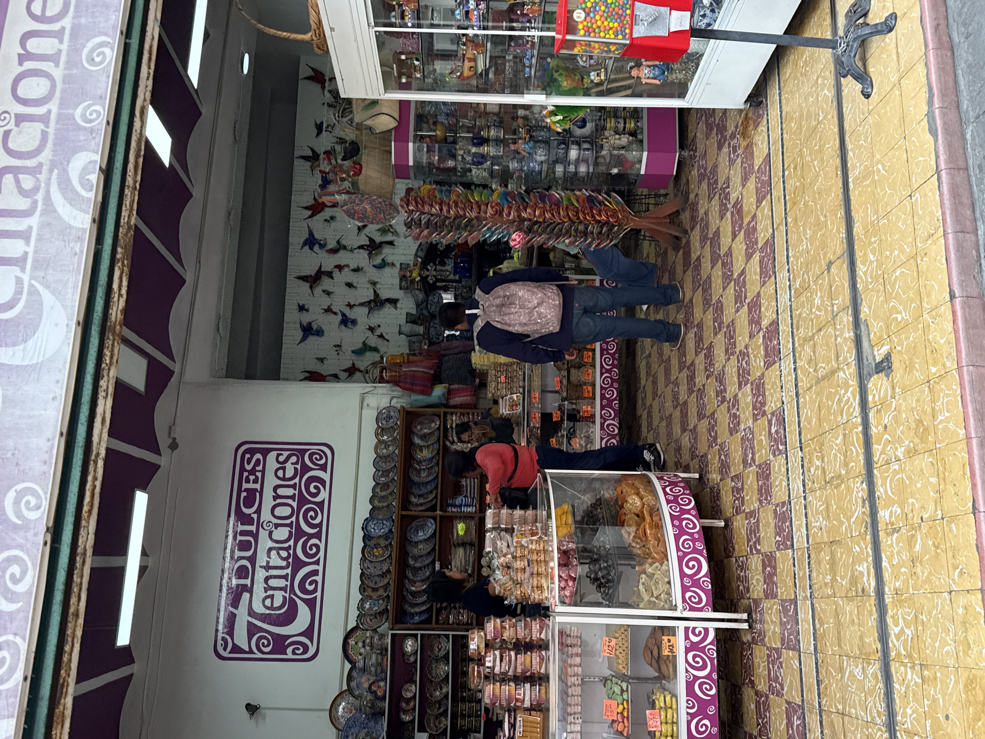 An image captures the entrance and interior of a vibrant shop named "DULCES Tentaciones" (Sweet Temptations) in Puebla, Mexico. The shop specializes in both traditional sweets and local crafts.
The interior features a checkered floor with brown and beige tiles. Along the left wall, decorative plates, likely ceramics, are neatly stacked on wooden shelves. Further back, numerous colorful paper or ceramic bird decorations (resembling swallows or hummingbirds) adorn a white wall.
In the foreground, on the right side of the shop entrance, a display case exhibits various dried fruits and candies, some with visible price tags like "$12.00" and "$13.00". Another glass display case towards the back holds more confectionery. Near the center, a large wooden rack displays an array of colorful, spiral lollipops.
Two individuals are visible inside the store. One person, wearing a dark blue jacket, jeans, and a light-colored backpack, stands near the lollipop display, facing away from the camera. Another person in a red top and dark pants is seen deeper inside the shop, appearing to browse items on shelves. At the far right, a gumball machine filled with colorful gumballs sits atop a counter.
The shop's name, "DULCES Tentaciones," is prominently displayed on a purple sign on the wall, featuring ornate white lettering and swirl decorations. The exterior sidewalk, visible on the right, is paved with yellow and white patterned tiles. A black ornate metal stand is positioned on the sidewalk. The bright ambient light suggests it is daytime.