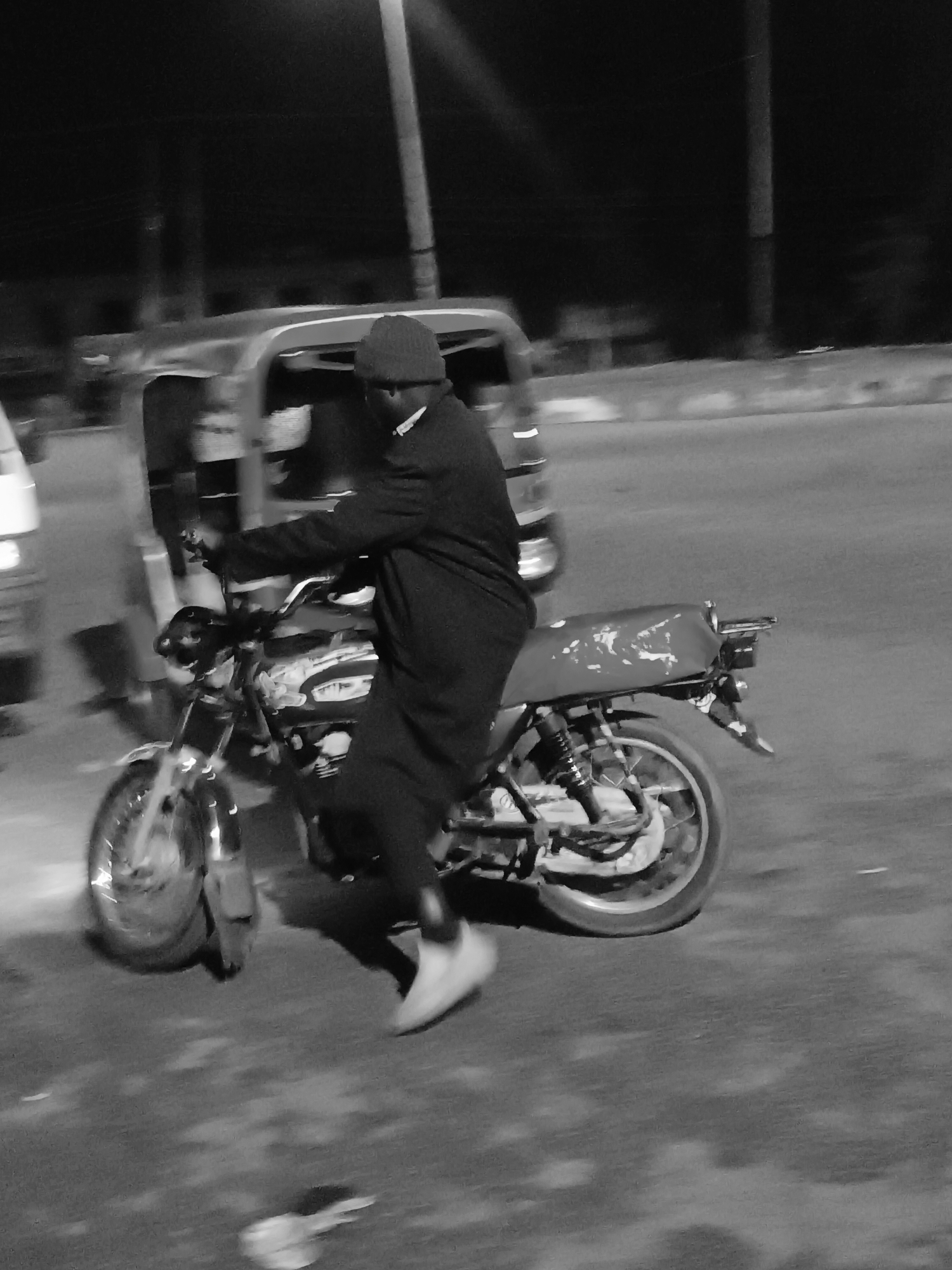A black and white photograph taken at night depicts a man riding a motorcycle on a road in what appears to be Bukuru, Nigeria. The man is wearing a dark overcoat, a knit cap, and light-colored sneakers. He is actively operating the motorcycle, which is moving, creating motion blur. Behind the motorcycle, an out-of-focus auto-rickshaw is also in motion. Street lights are visible in the background. The road surface is asphalt, and there are some indistinct objects or debris on the ground near the bottom of the frame.