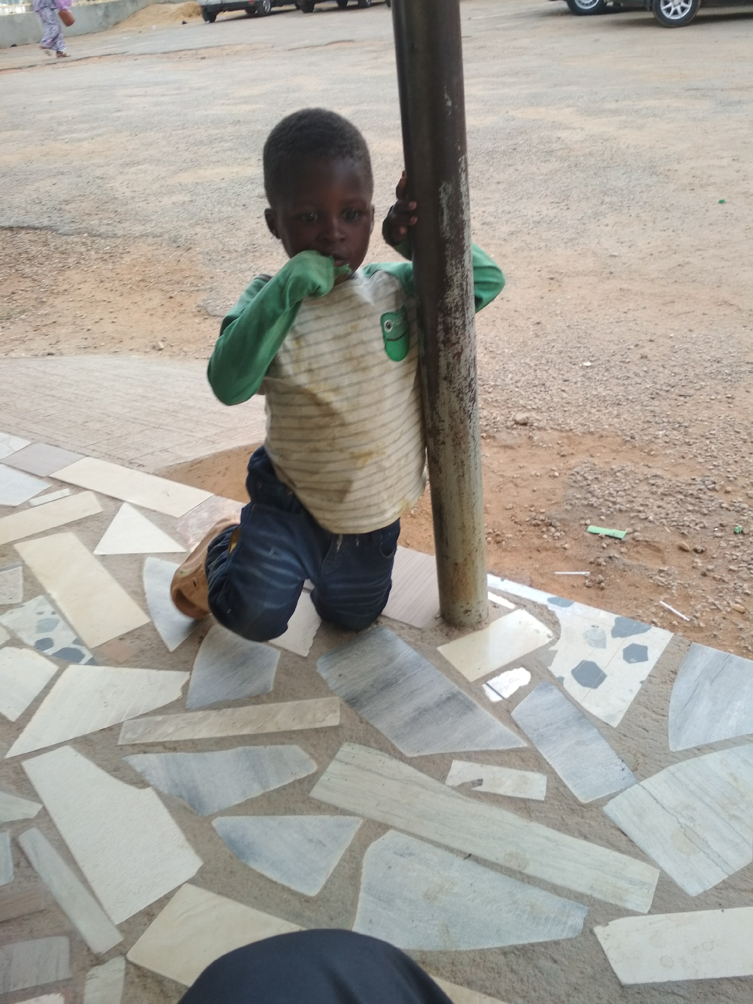 A young child kneels, grasping a metal pole with their left hand while their right hand is near their mouth. The child is wearing a long-sleeved shirt with green sleeves and a striped torso, along with dark-colored jeans and one brown sandal. The ground is tiled with irregular shapes and a rough, dirt surface extends beyond the tiled area, where several parked cars are visible in the background. The location is in a city of Unknown, Unknown.