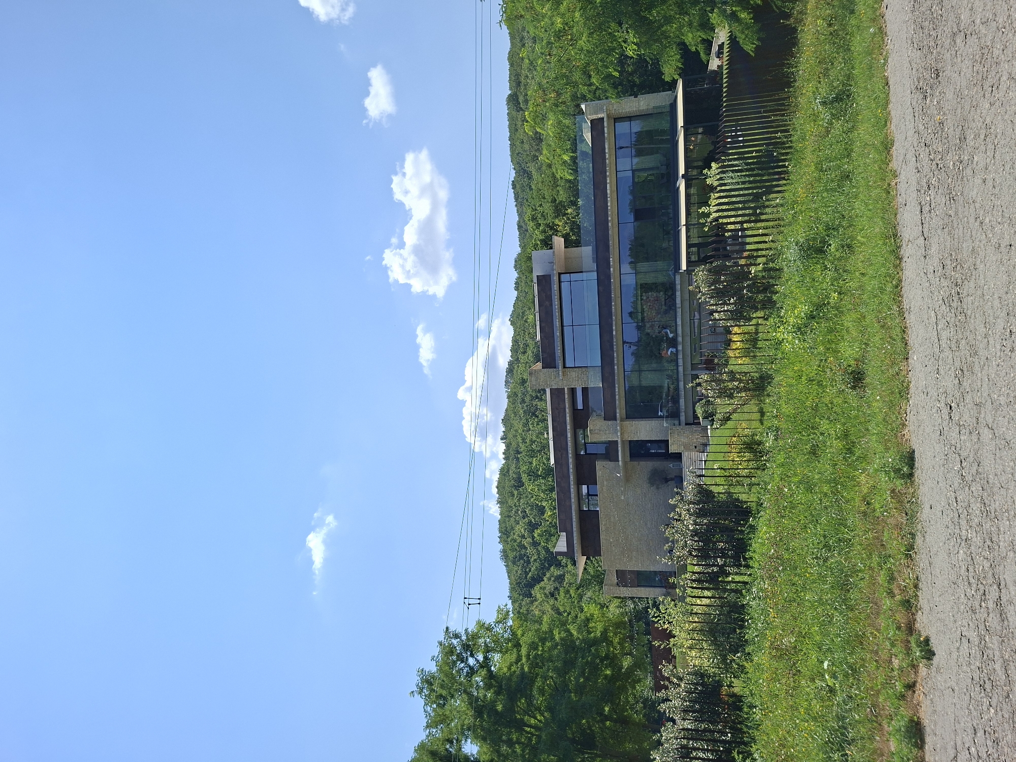 The image shows a modern, multi-story house with large glass windows and stone facade, situated on a green hillside covered in trees. The house is surrounded by lush vegetation and a black metal fence. Power lines are visible in the sky, stretching across the frame. In the foreground, there is a rough, gray road with patches of grass. The sky is blue with scattered white clouds. The scene appears to be a sunny day in a rural or semi-rural setting. This property is identified as being located in Condrița, Moldova. There are no people or animals clearly visible within the frame, though some indistinct figures may be seen through the glass windows of the house.