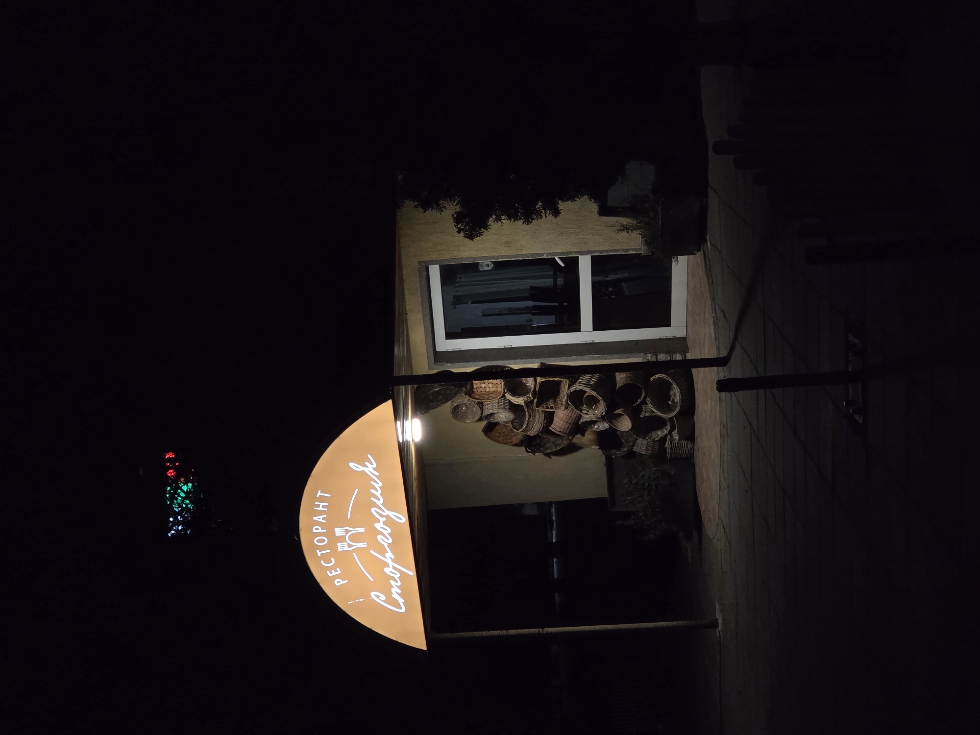 A dimly lit, slightly blurry image shows the exterior of the Стороводик restaurant in Pleven, Bulgaria, at night. A rounded, illuminated sign bearing the restaurant's name in Cyrillic ("Ресторант Стороводик") is prominently displayed. Behind the sign, numerous woven baskets are visible. The restaurant building is simple, with a visible window. A sidewalk and dimly lit objects are located to the right. Far to the left, a group of lights in red, green, and white are visible. The overall scene suggests a quiet, nighttime atmosphere.