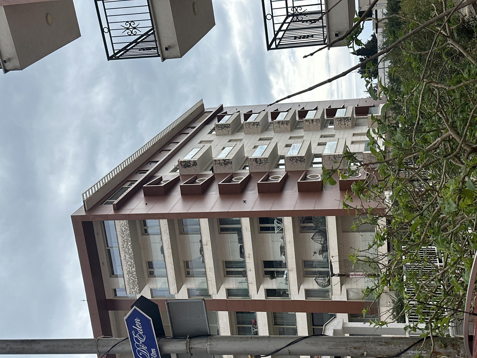 An outdoor image depicts a modern, multi-story residential building under an overcast sky, located in Budva, Montenegro, specifically indicated by a blue directional sign reading "Do Eden" and "50m" mounted on a grey utility pole in the left foreground. The main building features a light-colored facade with contrasting dark reddish-brown panels. Its left side exhibits recessed balconies with glass railings, while the right side presents protruding, box-like balconies with patterned sides and glass railings. Contents visible on some balconies include outdoor furniture such as tables, chairs, and two egg-shaped hanging chairs, along with potted plants. To the right of the frame, two light-colored concrete balconies with black wrought-iron railings, featuring decorative scrollwork, are partially visible from an adjacent structure. The immediate foreground is dominated by dense green foliage with some bare branches. No individuals are visible.