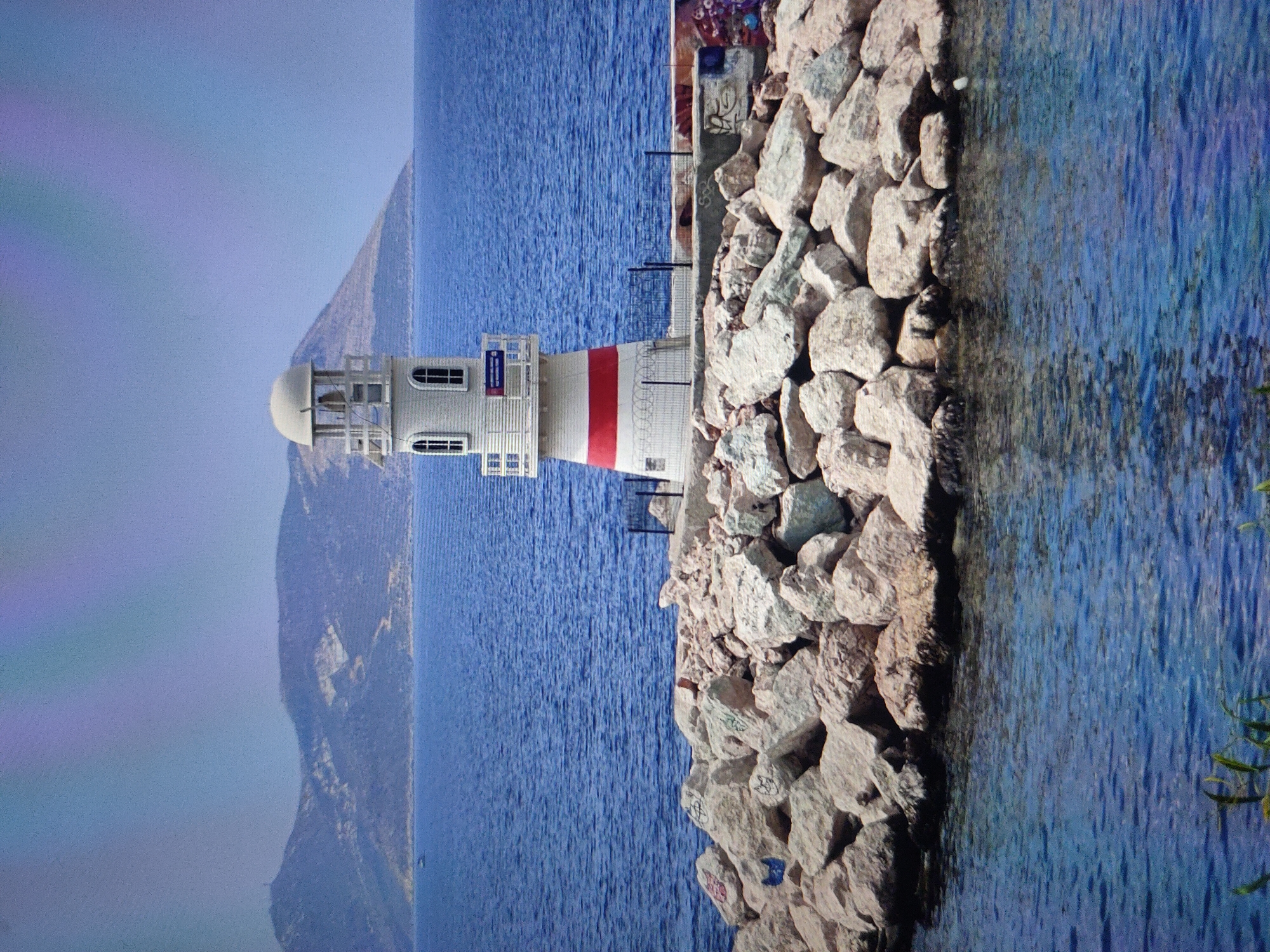 The image shows a lighthouse with red and white bands, situated on a rocky coastline. The lighthouse is white with arched windows and a cylindrical structure topped by a dome. A red band encircles the middle section of the lighthouse. The coastline is made of large, grey rocks, some of which are covered in graffiti. The sea is a deep blue and appears calm. In the background, a mountain range is visible under a clear blue sky. The image is oriented vertically.