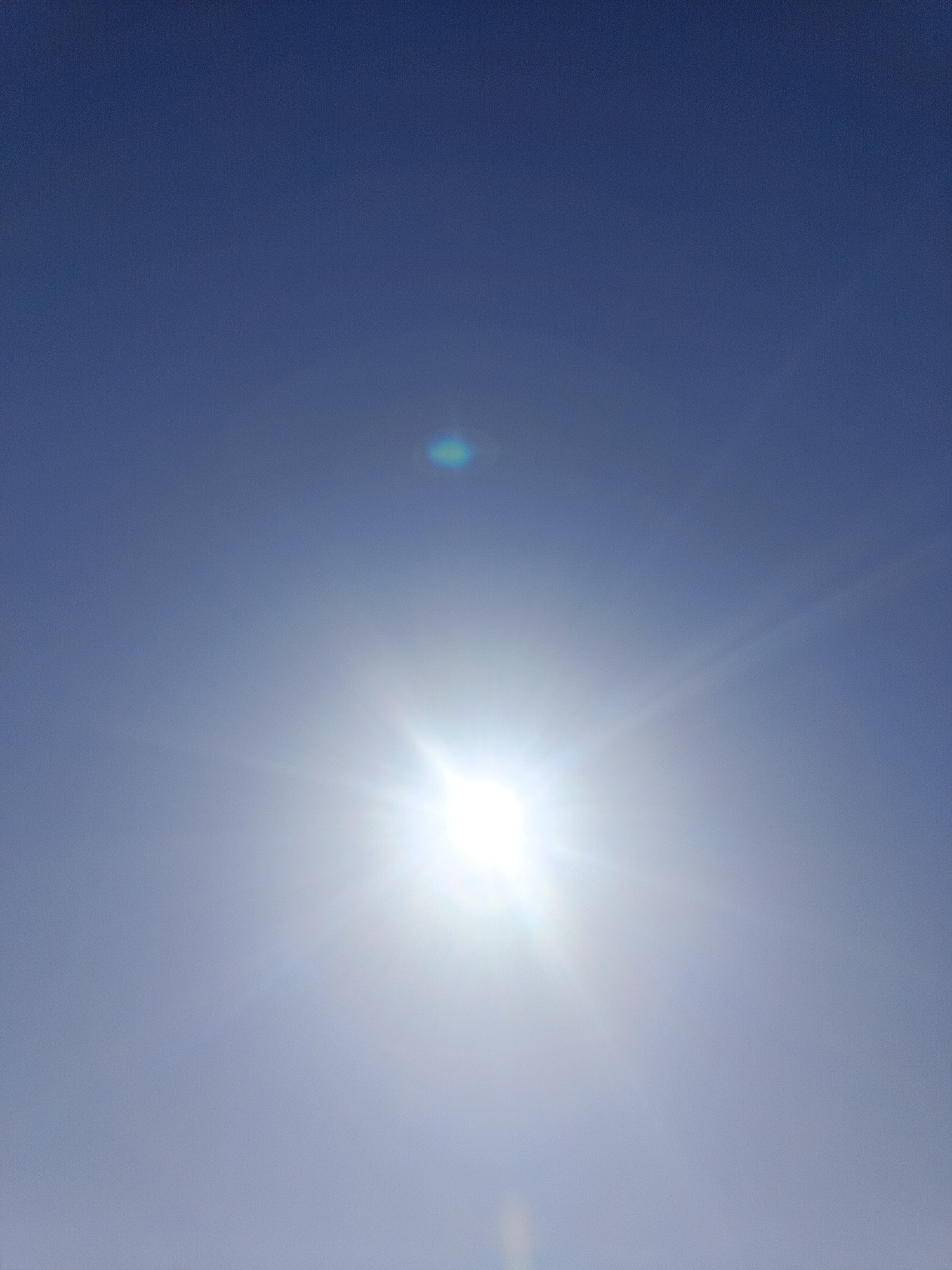 The image depicts a bright, clear sky. The sun is the dominant feature, appearing as a very bright, intense light source with rays emanating outwards. There is a faint circular rainbow-like artifact present in the sky. Based on the location information, this is likely a daytime scene in Chaikovsky, Russia, captured looking upwards towards the sky. There are no other discernible objects, people, or actions visible.