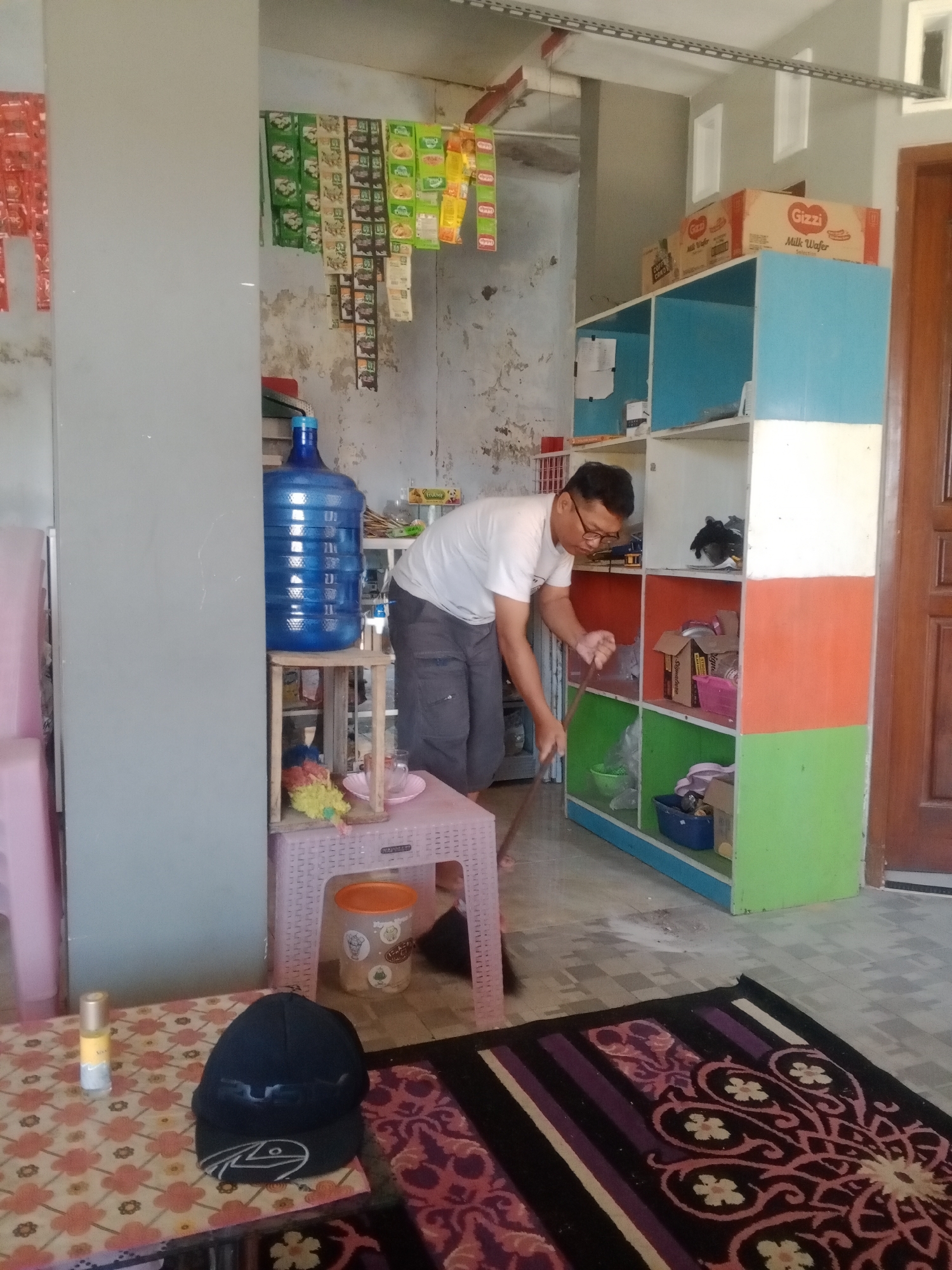 In Kemulan, Indonesia, a man wearing glasses and a white t-shirt is sweeping the floor with a broom in a cluttered room. A large blue water jug sits on a wooden stand next to a pink plastic stool with a cup and a flower on it. To the right, a colorful shelf unit holds various items, including boxes, bags, and containers. The room has a worn, textured wall and a tiled floor. A patterned rug is laid out in the foreground. The overall impression is one of a busy, lived-in space.