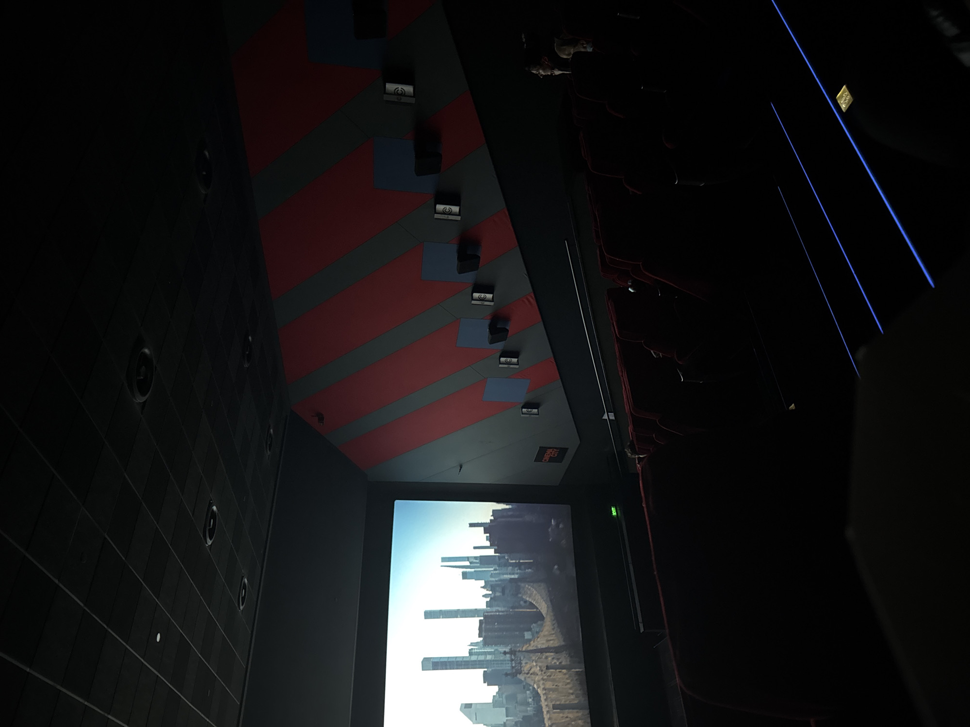 The image depicts the interior of a cinema, likely in Plovdiv, Bulgaria, with a large screen displaying a city skyline. The ceiling above the screen is patterned with alternating red and grey stripes, punctuated by speaker boxes. Seating is visible on the right side, appearing as red, tiered rows. A dark, tiled wall is on the left, with circular speaker-like elements spaced across its surface. The overall impression is that of a dark, indoor space focused on the projection screen.