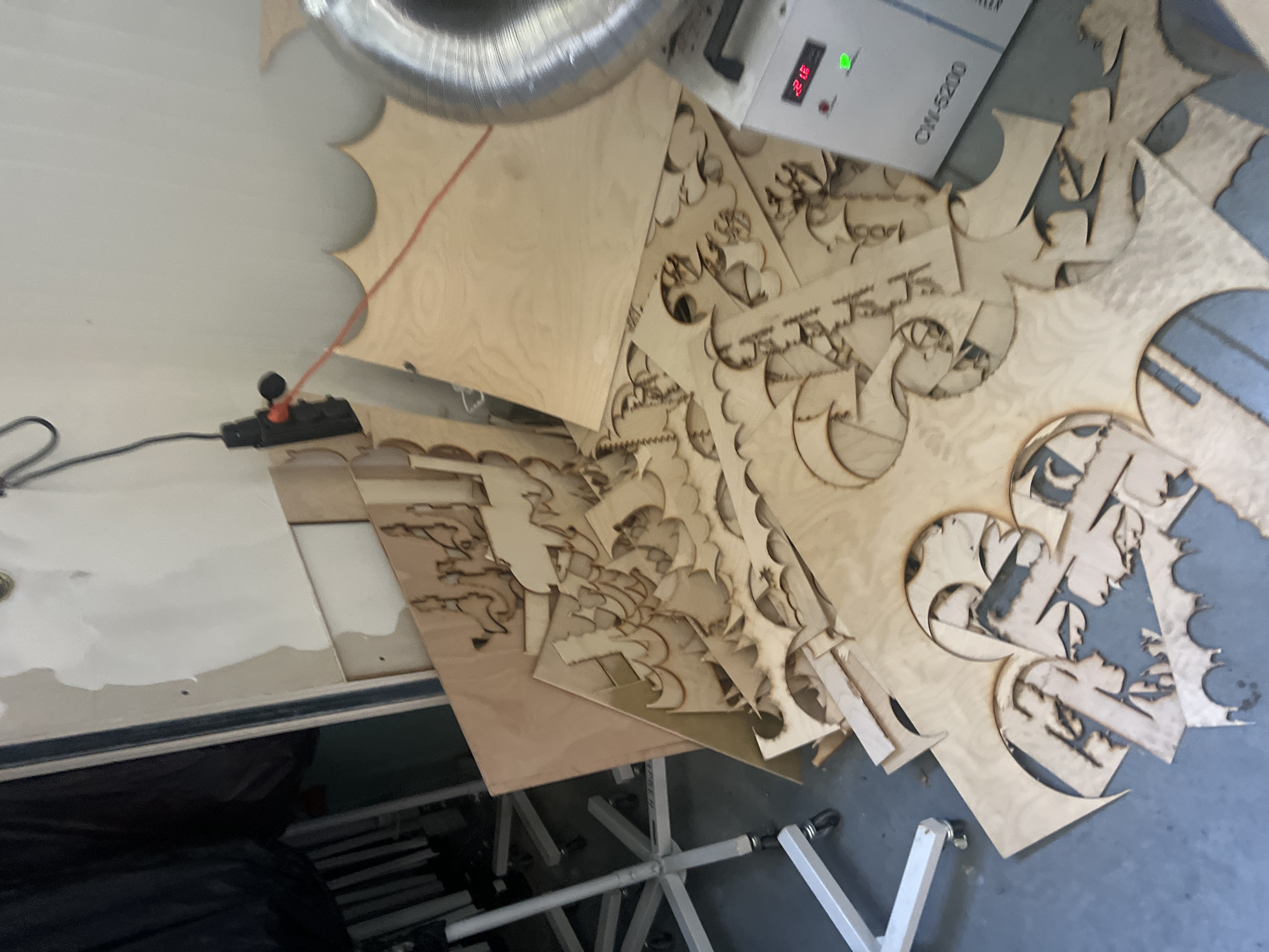 The image shows a cluttered workspace with various laser-cut wooden shapes scattered on a surface. The shapes appear to be decorative or artistic, featuring intricate patterns and designs. There's a CW-5200 chiller visible, along with an exhaust duct, suggesting the use of laser cutting equipment. The workspace seems to be a manufacturing or crafting environment. A black cable with a switch is also visible in the corner of the image. A black bag, and a metal trestle are visible at the lower left of the image. The location is not possible to determine from the image content.