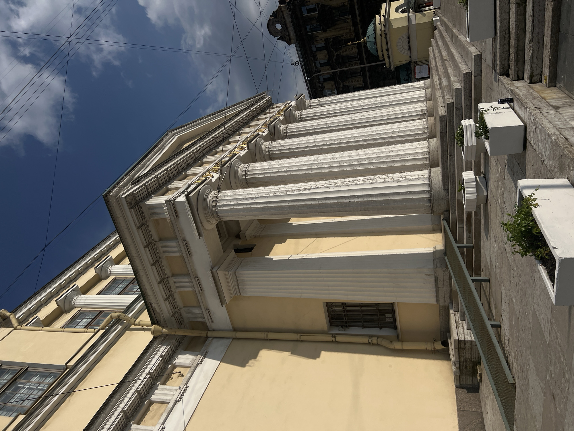 The image depicts the facade of a classical building in Saint Petersburg, Russia, characterized by its pale yellow walls and a prominent portico supported by fluted white columns. The building features ornate architectural details, including a decorative frieze with gold lettering and a pediment. In the foreground, a wide set of stone stairs leads up to the building's entrance, flanked by modern white planters containing greenery. A metal ramp is installed on the left side, providing accessibility to the steps. A drainpipe runs vertically along the yellow wall. In the background, other buildings are visible, including a darker, older structure with windows and a small domed building with a cross. Overhead, a network of power lines crisscrosses the blue sky, which is dotted with clouds. The scene is bathed in sunlight, casting shadows on the building's facade and the surrounding ground. There are no people visible in the image.