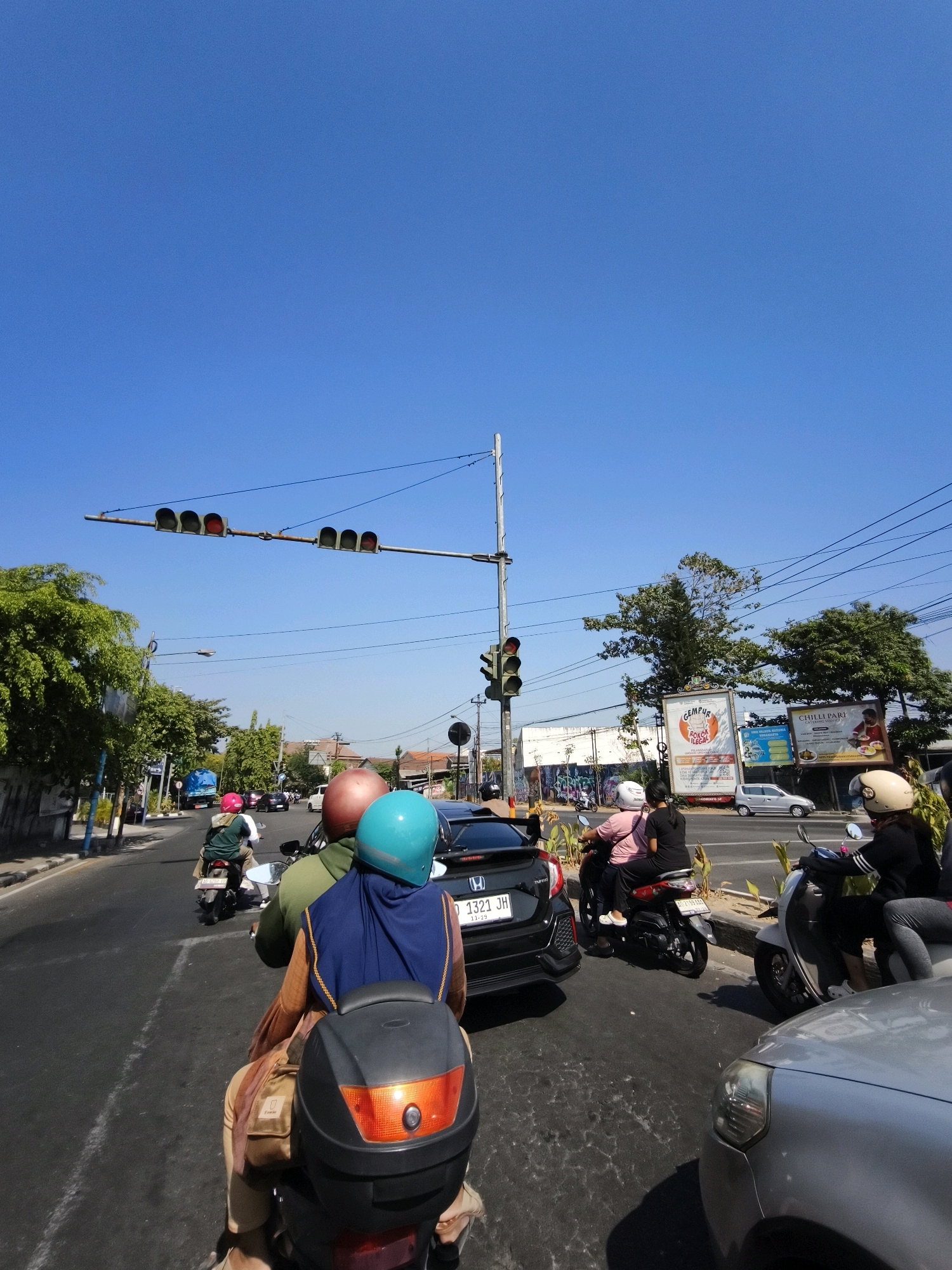 The image depicts a street scene in Surakarta, Indonesia, on a bright, clear day. The viewpoint is from the back of a motorcycle, looking forward. Several other motorcycles and a black car are also on the road, suggesting moderate traffic.
In the foreground, a person wearing a blue hijab and a tan outer garment is seated on a motorcycle, facing forward. To their left, another person with a brown helmet is also on a motorcycle. The motorcycle has a black storage box on the back.
Further ahead, on the left side of the road, a line of traffic lights shows red signals. A large green tree with dense foliage is visible along the roadside. On the right side of the road, a billboard with text in Indonesian, advertising "Gempur Rokok Ilegal," is displayed. Another billboard for "Chilli Pari" is also visible. A silver car is parked on the side of the road.
In the background, more trees and buildings line the street, with power lines crisscrossing overhead. The sky is a clear, vibrant blue. The overall impression is a typical urban street scene in Indonesia under a sunny sky.