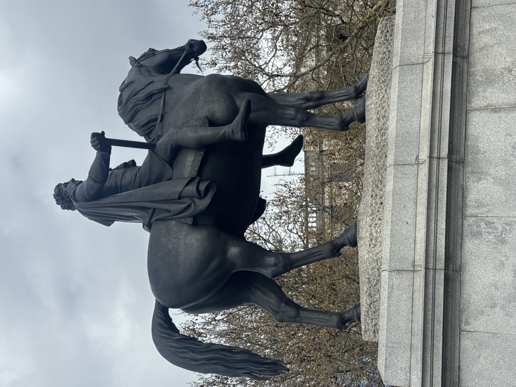 The image shows a statue of King Charles I on horseback, located in Trafalgar Square, London, United Kingdom. The statue is made of bronze and depicts the king in military attire, holding a baton. The horse is in a dynamic pose, with its front legs raised. The statue is mounted on a stone plinth. The background includes bare trees and a cloudy sky. A building is partially visible behind the trees.