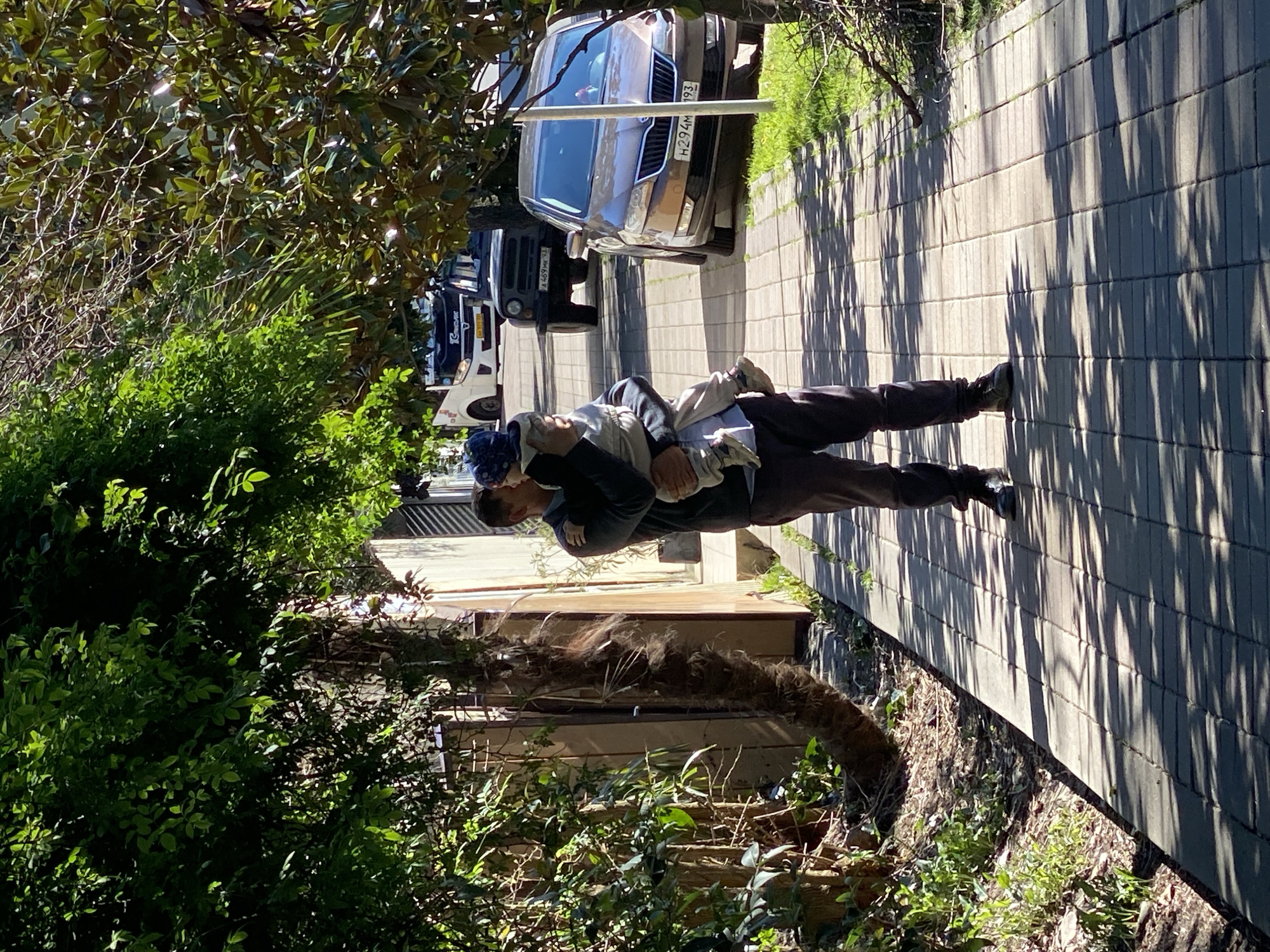 The image shows a man carrying a child on a paved sidewalk. The man is wearing dark pants, shoes, and a dark top. The child is wearing light-colored pants and a patterned hat. They are walking along a gray brick sidewalk, and several cars are parked alongside it. The license plate of one car reads "H294MP 193". There is greenery, including trees and bushes, on both sides of the sidewalk. The image appears to be taken in a residential area of Sochi, Russia.