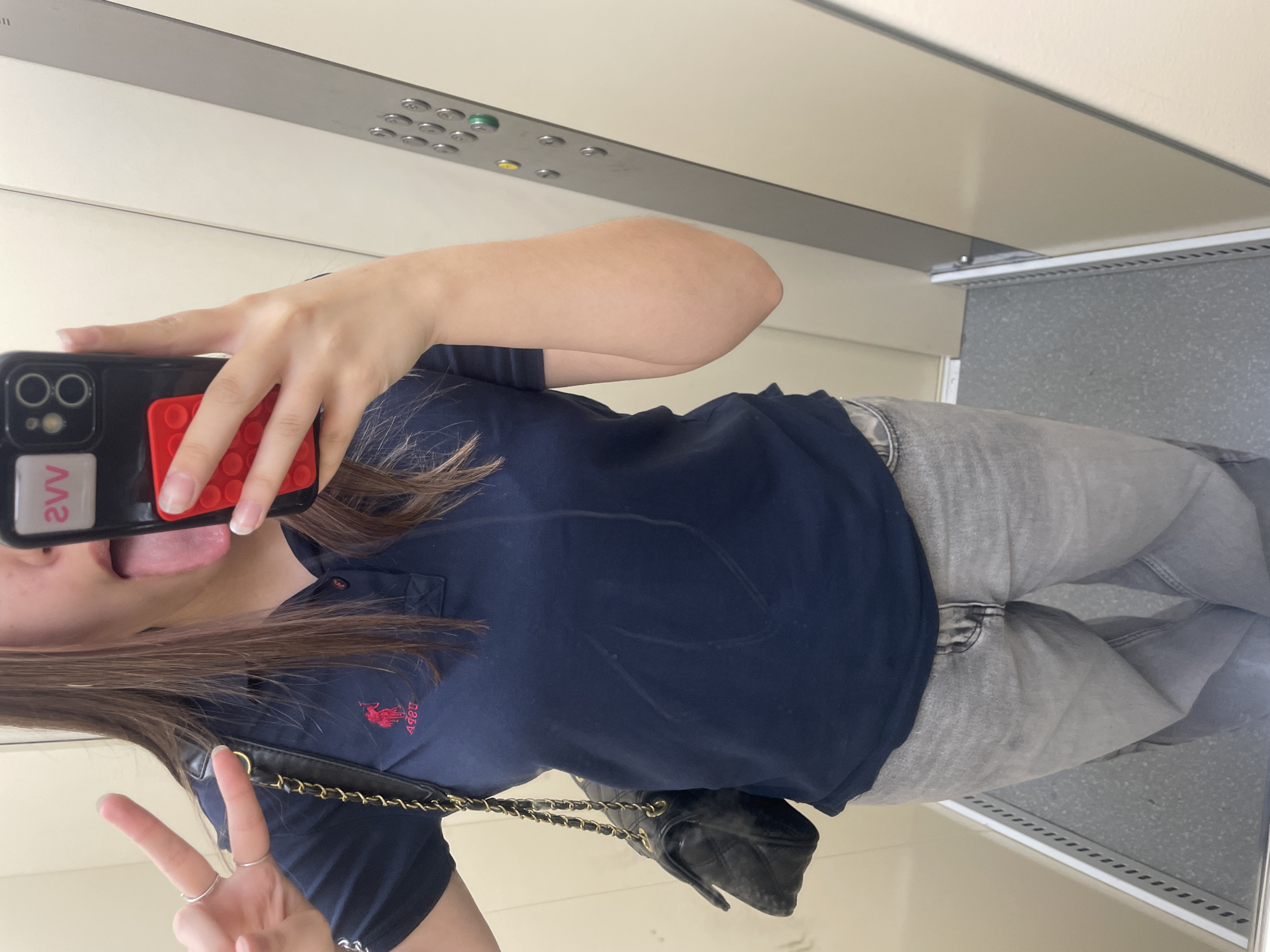 A person is captured in a mirror selfie, standing inside what appears to be an elevator. The individual, seen from the waist up, is wearing a navy blue short-sleeved polo shirt with a red logo, likely "U.S. POLO ASSN." with a horse and rider motif, and light grey jeans. They are holding a black smartphone with a red textured pop-it accessory and a white sticker or card on the back displaying the mirrored text "2VV" (originally "VV2"). The person's tongue is playfully extended.
On their other hand, adorned with several simple silver rings, they are making a peace sign. A black quilted shoulder bag with a gold chain strap is visible, hanging from their shoulder. The background, a reflection in the mirror, shows the interior of an elevator with a control panel featuring numerous buttons, some illuminated with green and yellow lights. The elevator walls are a light, neutral color, and the floor is speckled grey. The lighting is artificial and consistent with an indoor setting.
