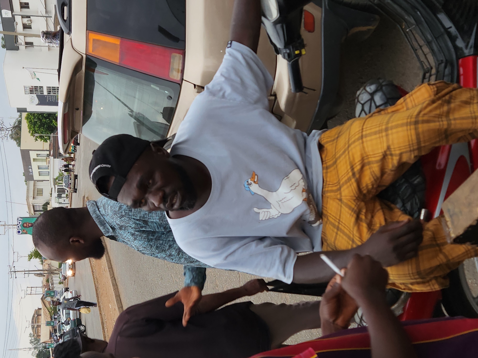 The image shows three men interacting on a street in Jos, Nigeria. One man is sitting on a red motorcycle, wearing a light blue t-shirt with a goose design and yellow plaid pants. He's holding a white object in his right hand and appears to be engaged in conversation. Another man, wearing a patterned blue shirt, stands beside the motorcycle, seemingly talking to the seated man. The third man is in the foreground with a brown shirt. A car is partially visible behind the motorcycle. The street is lined with buildings and utility poles, suggesting an urban setting. A billboard is located in the background. Cars are parked along the street.