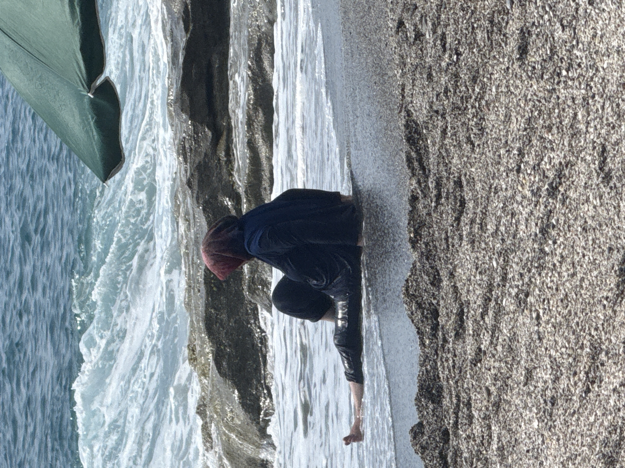 A woman wearing a dark blue full-body swimsuit and a red headscarf sits on the wet sand at the edge of the sea in Alanya, Turkey. Her legs are extended into the shallow water, and waves are gently lapping around them. Behind her, dark rocks emerge from the water, and further out, the turquoise sea stretches to the horizon. A portion of a green beach umbrella is visible in the upper right corner of the frame, casting a shadow over the scene. The foreground is covered in small, dark pebbles.