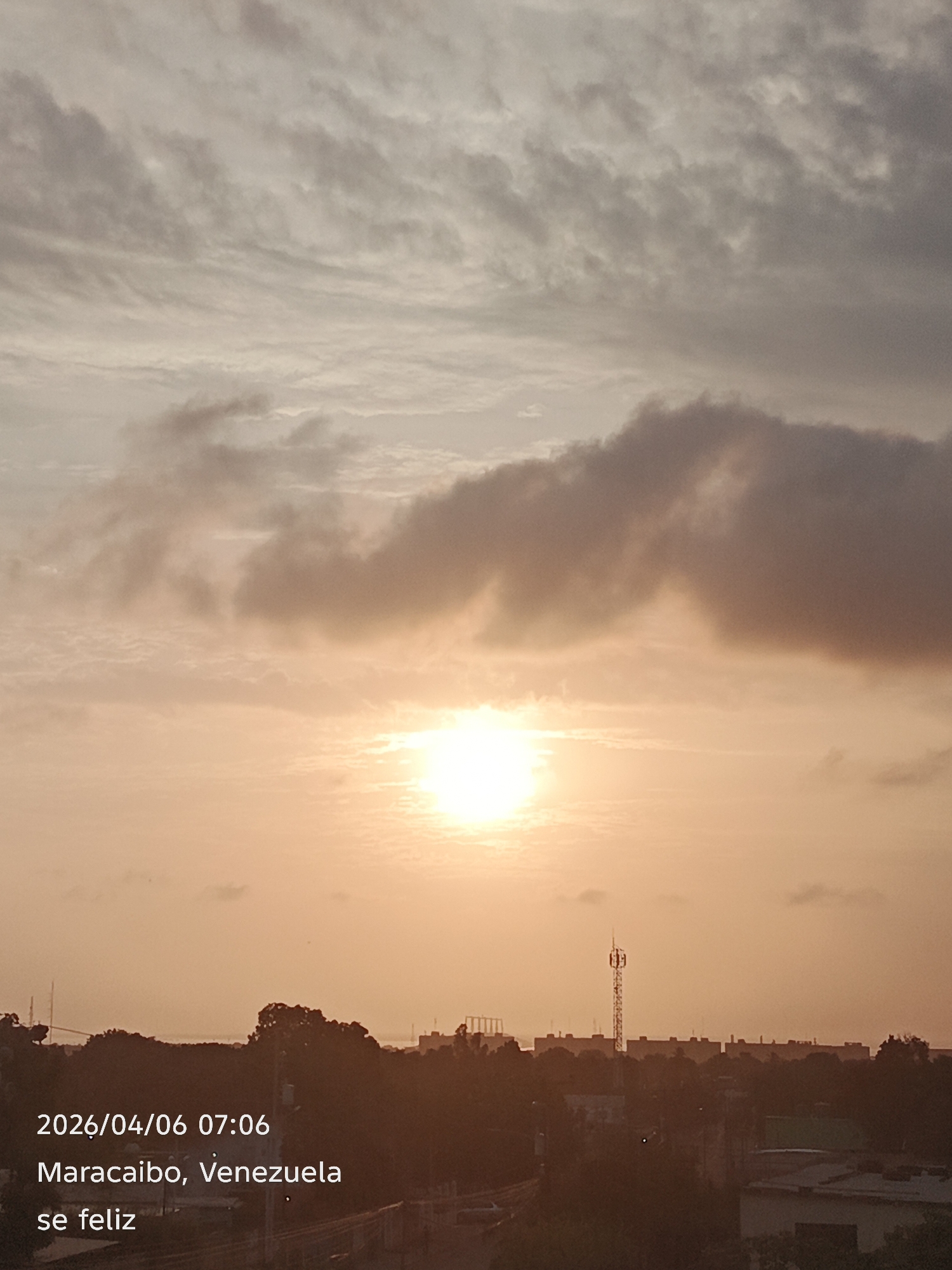 The image captures a sunrise or sunset in Maracaibo, Venezuela, on April 6, 2026, at 07:06 AM. The sky is filled with clouds, and the sun is shining brightly through them. In the foreground, there is a silhouetted skyline of trees and buildings, including a cell tower. The text "se feliz" (be happy) is displayed in the lower left corner.