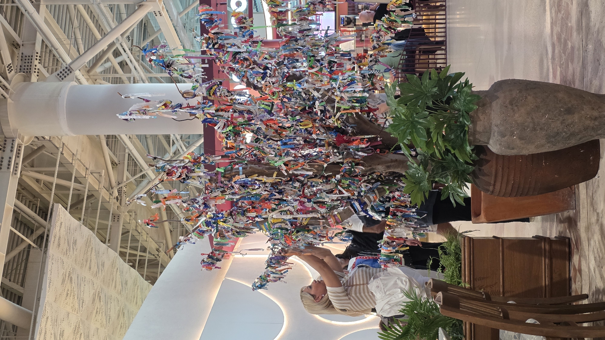 An indoor photograph captures a vibrant tree-like art installation adorned with numerous colorful tags or ribbons. The artwork is set within a spacious public area, possibly an airport or shopping mall, characterized by a high ceiling supported by a network of white metal beams. A woman, identified by her blonde hair and light-colored striped shirt, stands near the artwork and appears to be adding or adjusting one of the tags. A large, aged terracotta pot with a plant inside is positioned beside the art piece. The location is Unknown, Unknown.