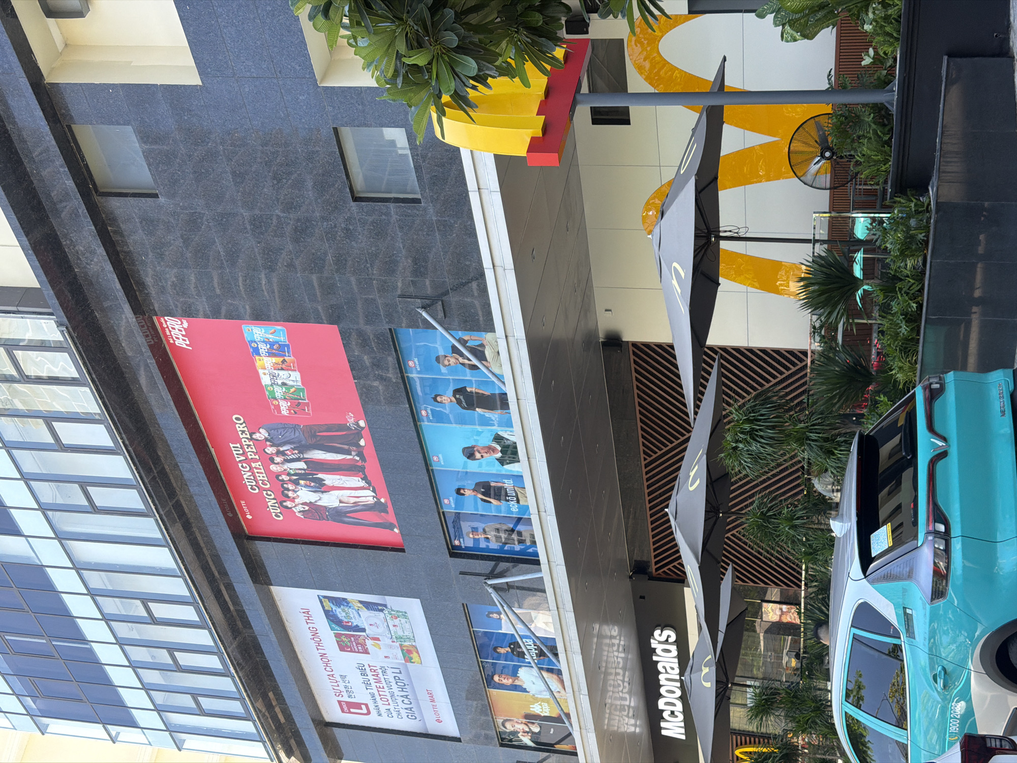 The image shows the exterior of a McDonald's restaurant in Nha Trang Ward, Vietnam, likely on a sunny day. The restaurant is located on the ground floor of a modern, multi-story building with a dark granite facade and large glass windows. A prominent McDonald's sign with the iconic golden arches is visible. In front of the restaurant, there are outdoor seating areas with grey umbrellas and potted plants. A bright turquoise car with "VinFast" branding is parked on the left side of the image. Above the McDonald's entrance, large advertisements are displayed on the building's facade. One advertisement is for Lotte Mart and features Korean text and product images. Another is a large red billboard with text in Vietnamese, "Cùng vui cùng chia Pepero" which translates to "Let's have fun, let's share Pepero," featuring a group of people and colorful Pepero snack boxes. Several framed posters of young men are also visible on the building, likely promotional images for various brands. The overall scene depicts a commercial area with a mix of retail and fast-food establishments.