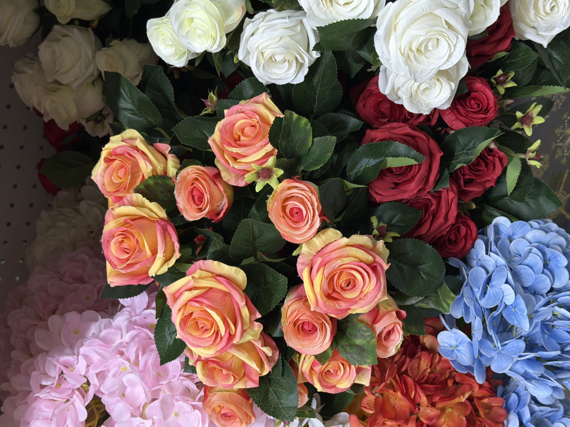 The image shows a close-up of various artificial flowers. Predominantly, there are peach-colored roses with yellow edges, along with clusters of white roses, red roses, pink hydrangeas, and blue hydrangeas. Green leaves are visible among the flowers. The setting appears to be indoors, possibly a store or display, but the precise location within Perm, Russia, cannot be determined from the image alone.