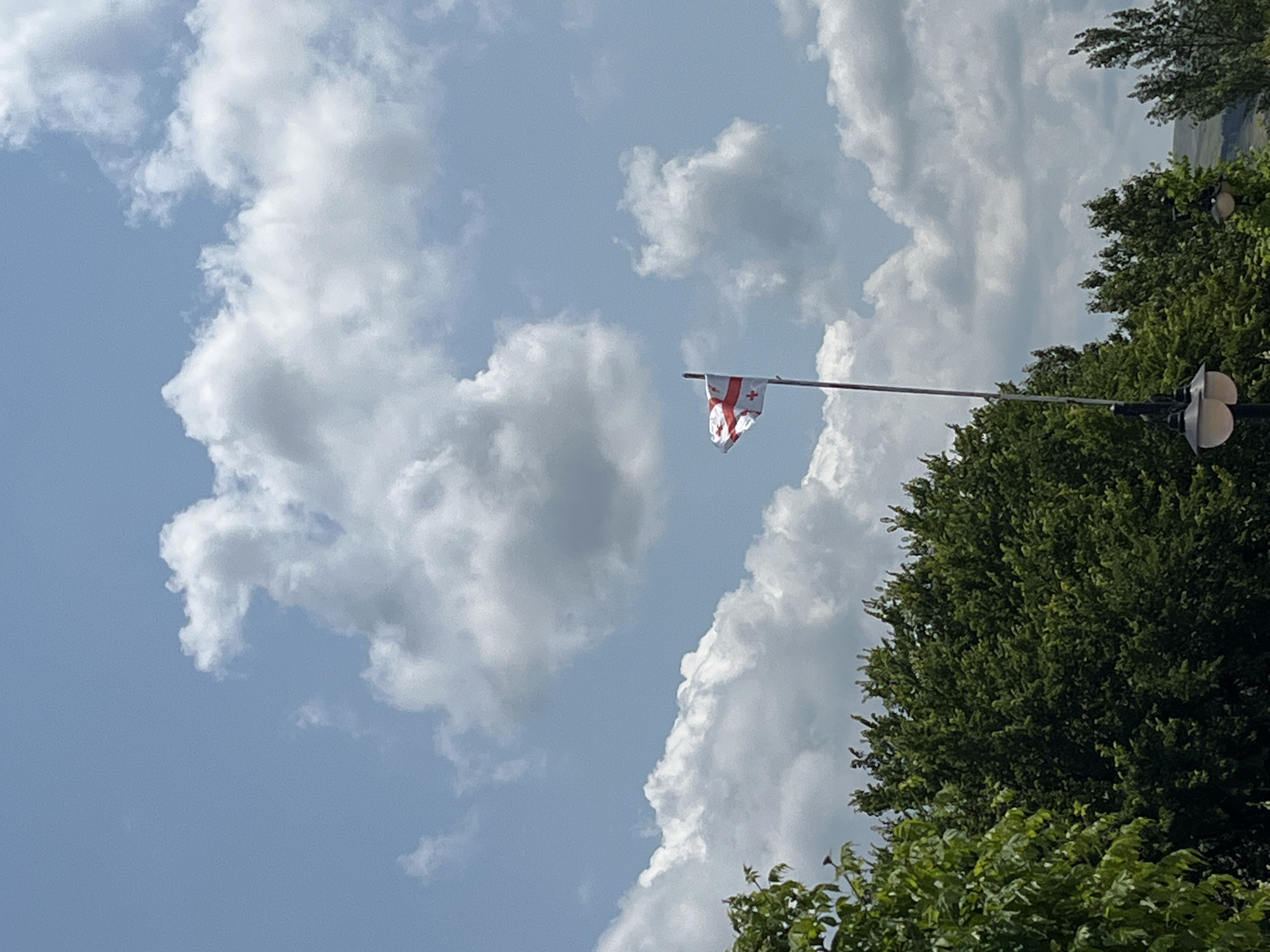 The image shows a flag with a red cross and four smaller red crosses on a white background, likely the flag of Georgia, fluttering on a flagpole against a bright blue sky with white, fluffy clouds. The bottom of the image is partially filled with dark green trees, suggesting an outdoor setting. There are also streetlights visible in the foreground. The scene appears to be captured during the daytime under clear weather conditions. Based on the flag and vegetation, this scene could be located in Tbilisi, Georgia.