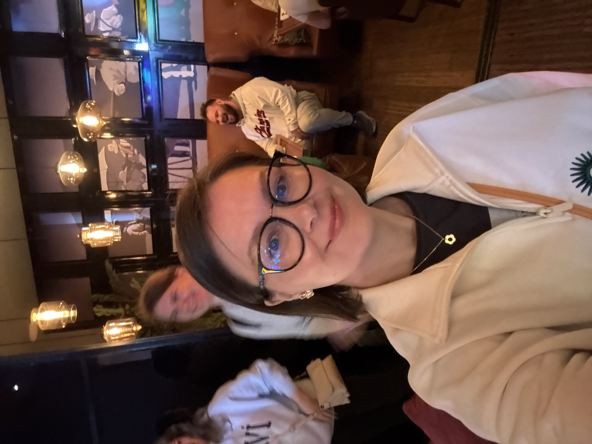 The image is a selfie taken indoors, likely in a cafe or restaurant. A woman with glasses is in the foreground, smiling slightly. Behind her, other people are visible, one of whom is a man reclining on a leather couch, and another a woman. The background features a wall with a series of framed pictures illuminated by decorative light fixtures. A clue to the location being Izhevsk, Russia, would depend on unique identifiable details within the image – such as a specific brand, logo, or architectural feature known to exist exclusively in that city. Without such details, it is not possible to confirm the location definitively.