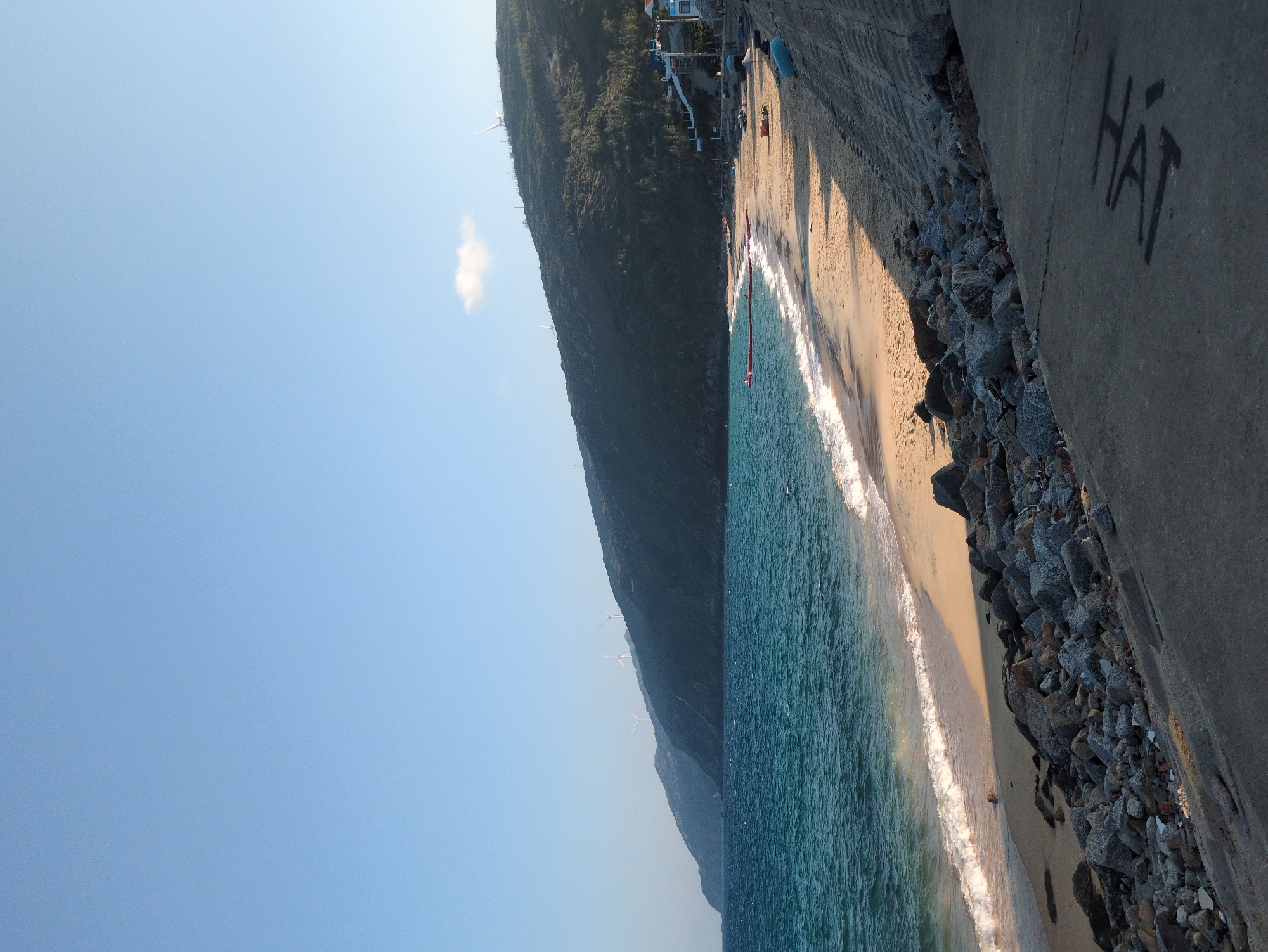 The image depicts a coastal scene in East Quy Nhon Ward, Vietnam, under a clear, light blue sky with a single small white cloud. The scene is dominated by a stretch of sandy beach curving into the distance, bordered by the turquoise-blue ocean on the left and a steep, verdant, tree-covered hillside on the right and in the background.
Gentle white waves break along the shoreline. In the water on the right, a red buoy line extends from the beach, with one individual visible standing near its end. On the sandy beach itself, several small, dark-colored boats or canoes are resting near the water's edge.
The hillside in the background is dotted with multiple white wind turbines along its ridges. Towards the base of the hill on the far right, a cluster of residential structures, featuring white and blue elements, is visible, accompanied by a pathway or stairs leading upwards.
In the immediate foreground, a grey concrete path or sea wall slopes downwards, partially obscured by shadows. Dark graffiti resembling the characters "LA HI" is present on its surface. Between this concrete structure and the sandy beach, a significant accumulation of grey rocks and miscellaneous debris forms a rough embankment. The light suggests a late afternoon setting, casting long shadows.