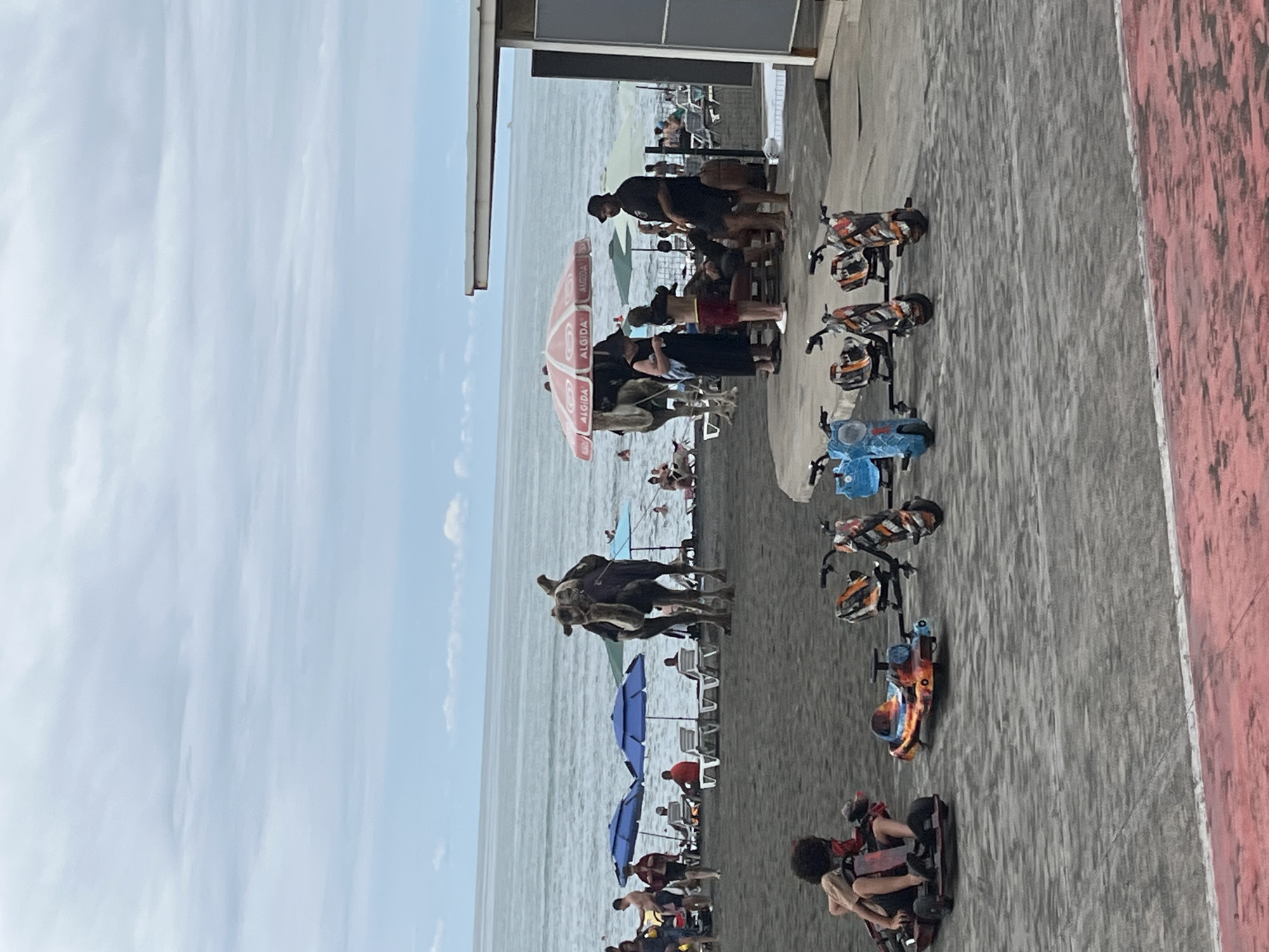 The image depicts a busy beach scene in Ureki, Georgia, a town known for its magnetic black sand beaches. In the foreground, several colorful electric drift trikes are parked on the sand, suggesting an amusement area. A child is seated on one of the trikes on the far left, looking towards the right.
Further back, more beachgoers are visible. Two camels stand on the sand, with people gathered around them, possibly for rides or photos. Several blue beach umbrellas are set up, providing shade for lounge chairs where some individuals are relaxing. The sea is in the background, with gentle waves and a cloudy sky overhead. A white building, likely a beachside establishment, is visible on the right side of the frame, extending partially into the image. The overall atmosphere is one of a lively tourist destination.