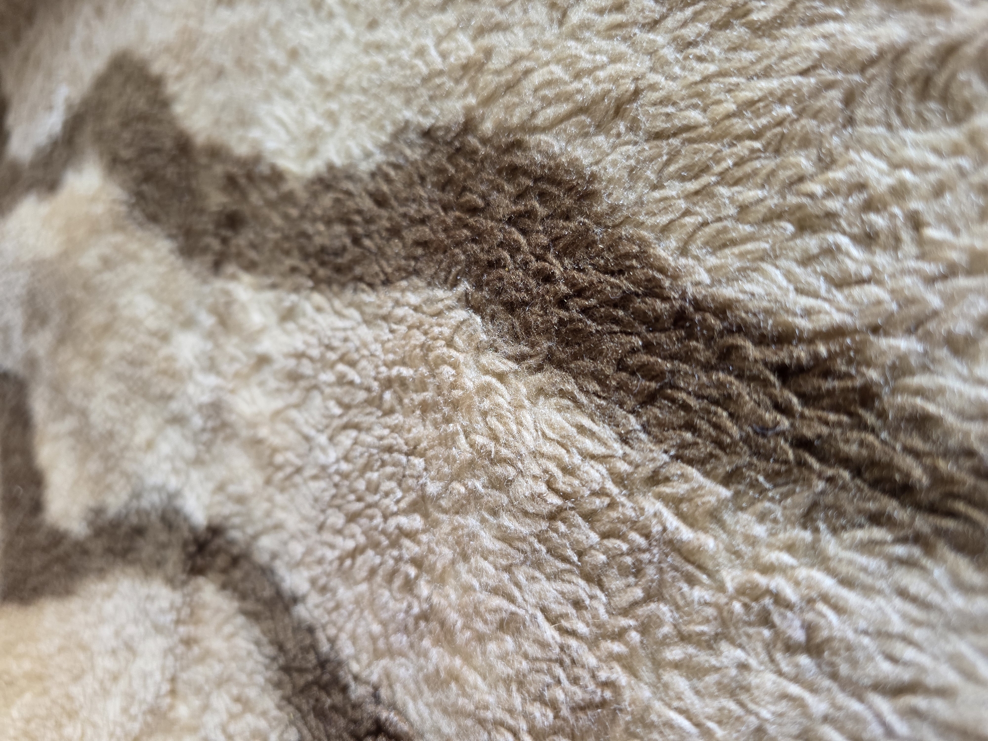 The image is a close-up, shallow depth of field shot of a textured, plush fabric. The fabric has a pattern of irregular, wavy bands of light beige and medium brown. The texture of the fabric appears soft and fibrous, with visible individual fibers creating a fluffy appearance. The lighting suggests a natural, diffused source, highlighting the raised areas of the texture. The image is a still life of the fabric itself and does not contain any elements that would suggest a location in Kyiv, Ukraine, or any other specific environment, people, or actions.