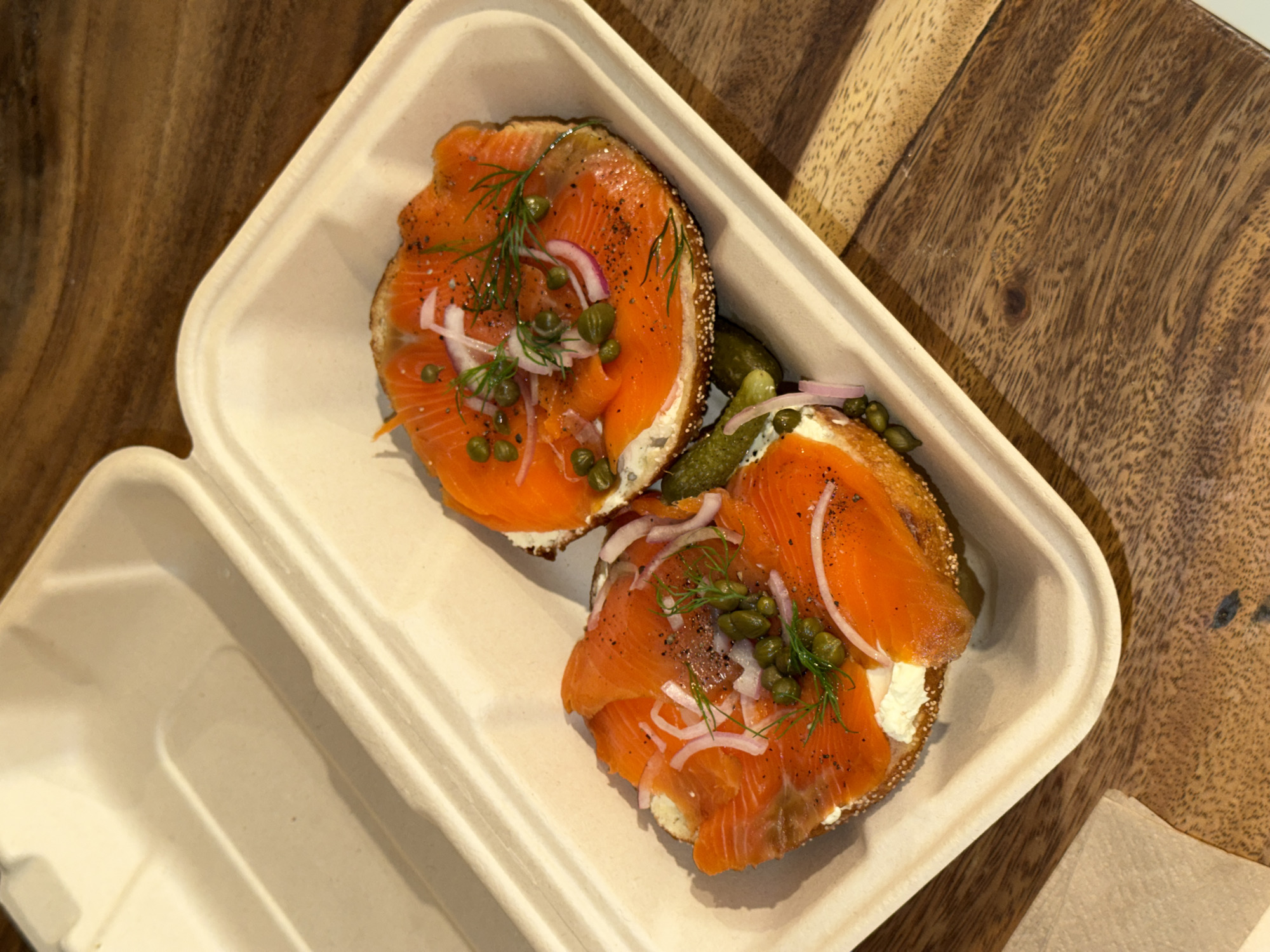 An overhead shot captures two halves of an open-faced bagel sandwich, artfully arranged in a light beige, possibly biodegradable, food container. Each bagel half is generously topped with layers of vibrant orange smoked salmon (lox) over a spread of white cream cheese. The salmon is garnished with small, dark green capers, thinly sliced purple-red onions, and fresh green dill sprigs, with black pepper visibly sprinkled over the top.
The food container, with its textured surface, occupies the right and central portion of the frame, with the left compartment of the container empty. It rests on a warm-toned, rich brown wooden surface, possibly a table or serving board, which fills the background with its distinct grain patterns and knots. A lighter wooden element is partially visible in the upper right corner. The overall scene suggests a freshly prepared meal, likely breakfast or brunch, presented in a casual setting. There is no visible text in the image. Based on the provided context, this scene is located in North Kuta, Indonesia.