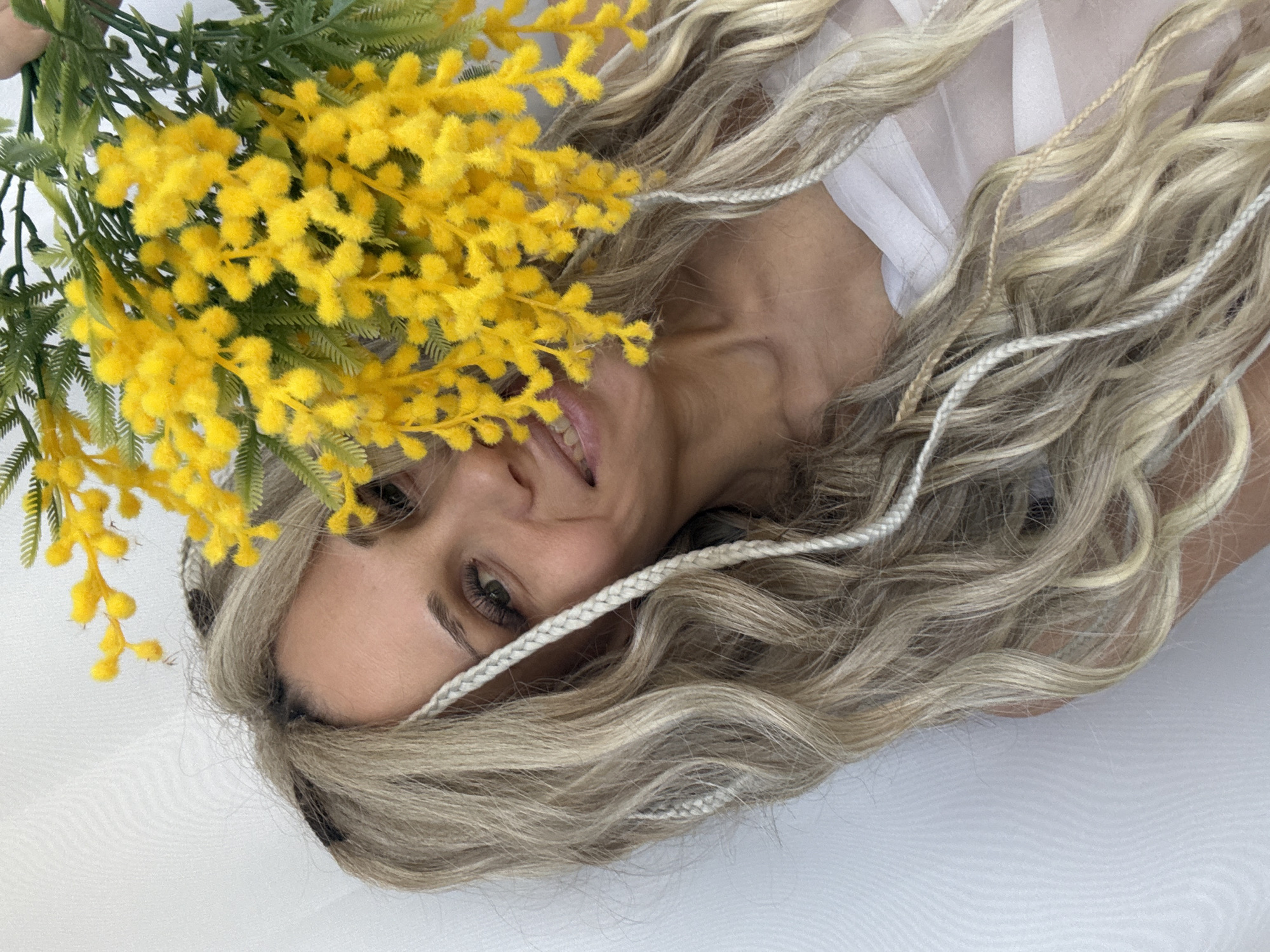 A woman with long, blonde hair and braids is lying on a white surface. A cluster of bright yellow flowers with green leaves partially covers her face. The location appears to be indoors.