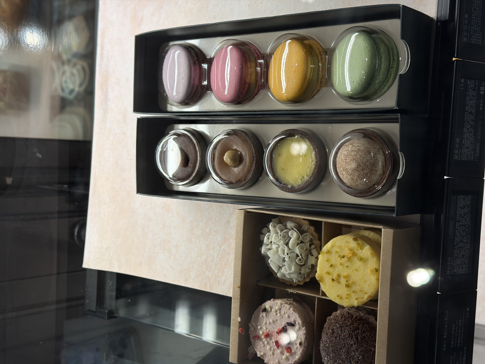 This is a still life photograph taken in Chongqing High-tech Zone, China. The scene appears to be a display case, potentially in a bakery or confectionery shop, showcasing various desserts. The focus is on three open boxes of sweets.
The top two boxes contain macarons. The top box has four macarons in a row, each of a different color: light purple, pink, yellow, and green. Each macaron is in its own clear plastic bubble insert. The box below contains four filled tartlets, also in individual clear plastic bubble inserts. They appear to be filled with chocolate cream, brown nut-flavored cream, a bright yellow custard, and another cream with flecks, all in individual clear containers.
The third box contains cookies or small pastries arranged in a 2x2 grid. The pastries appear to be a light colored tart, a shortbread cookie speckled with yellow and green sprinkles, a pastry with pink frosting, and a chocolate-covered dessert.
To the right of the boxes, several black boxes with Chinese characters are stacked vertically, possibly containing labels, price, or product information.
The image is brightly lit and appears to be taken at a slight angle. The background behind the display case is partially visible through the glass, showing a blurred environment.