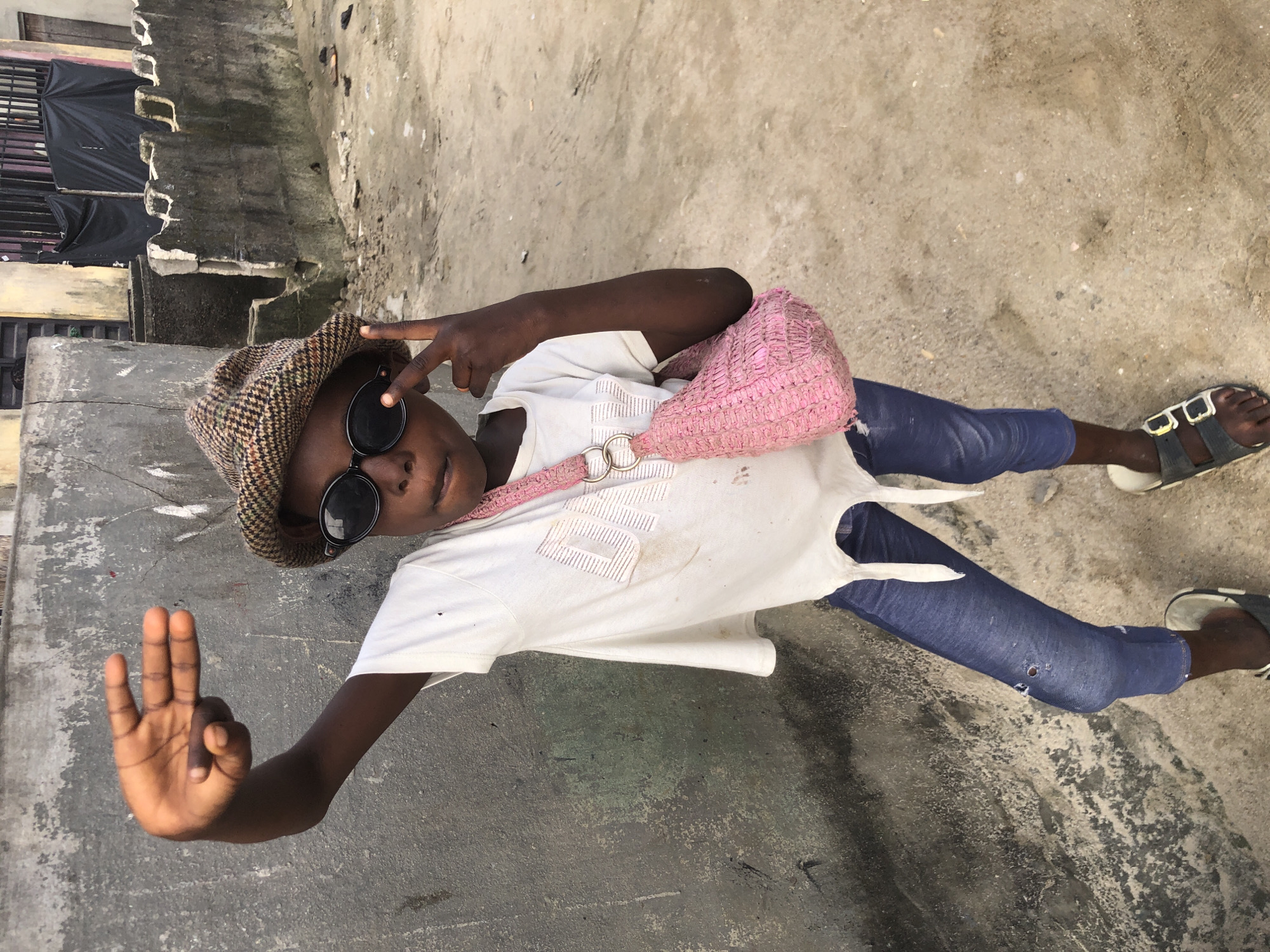 A young person stands in a dusty urban area, possibly Ado, Nigeria, with their left arm raised. They are wearing a plaid cap, round black sunglasses, and a white t-shirt with a pink woven purse slung over their right shoulder. The t-shirt has a frayed hem. They are also wearing blue jeans that appear to be ripped at the knee and dark sandals. The person is making a gesture with their right hand, with their index and middle fingers extended. The background consists of a concrete wall and a dirt or sandy ground. There are some buildings and structures visible in the distance. The lighting suggests it is daytime.