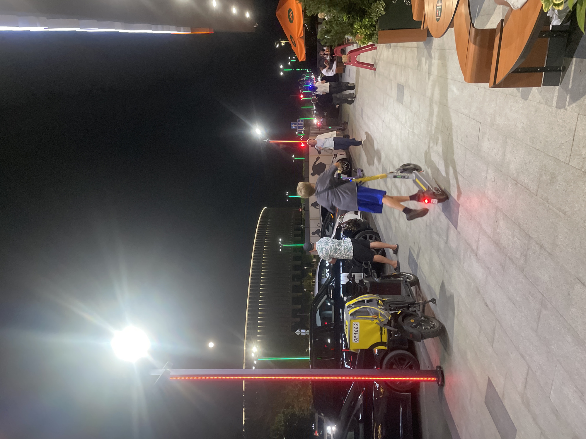 In Krasnodar, Russia, an urban scene unfolds at night under a bright streetlight. The foreground features a person riding a yellow and silver electric scooter, their legs extended. Adjacent to the sidewalk, a black electric scooter with a yellow cargo box, displaying the license plate "OP1682," is parked. Further back, several parked cars, including a dark-colored SUV, are visible. A large, modern building with a distinctive facade and illuminated upper sections dominates the background. Numerous streetlights, some emitting green light and others red, line the street. People are seen walking along the sidewalk and seated at outdoor tables, suggesting a public gathering area.