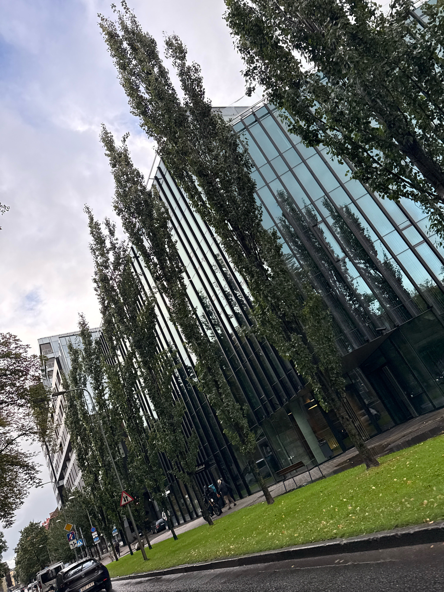 The image depicts a street scene in Tallinn, Estonia, focusing on a modern glass-fronted building with a row of tall, slender trees lining the sidewalk. The building's facade is highly reflective, mirroring the sky and the surrounding trees. The trees are densely leafed, suggesting it is either late spring or summer. A green lawn runs alongside the building, interspersed with more trees. The street in the foreground appears wet, possibly from recent rain, with a black car visible. Several traffic signs are present on the sidewalk. A few individuals are walking on the sidewalk, and some are on a scooter or motorcycle. The sky is overcast with some patches of lighter clouds. The overall impression is an urban setting with contemporary architecture integrated with a significant amount of greenery.