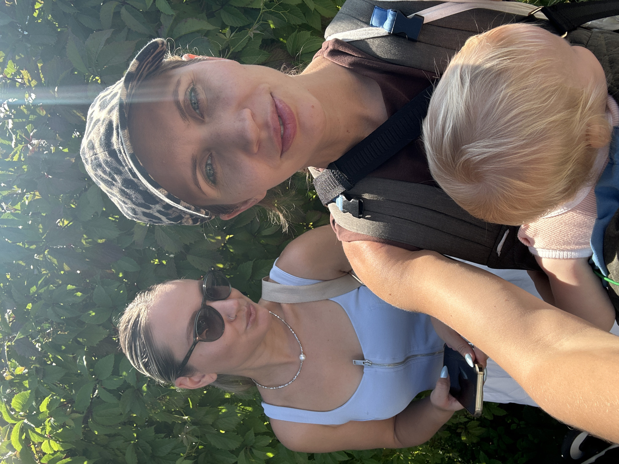 The image depicts two women and a baby outdoors, in front of a dense green leafy background that appears to be foliage. The woman in the foreground, wearing a leopard-print baseball cap and a brown shirt, is holding a baby in a carrier. She is looking towards the camera with a slight smile. The baby, with blonde hair, is facing away from the camera. To the left, a second woman, wearing a light blue sleeveless top and sunglasses, is holding a smartphone. She is looking down at the phone. The setting appears to be a sunny day, with light filtering through the leaves, suggesting it might be a park or a garden. The location is not specified within the image itself and cannot be determined to be Vsevolozhsk, Russia.