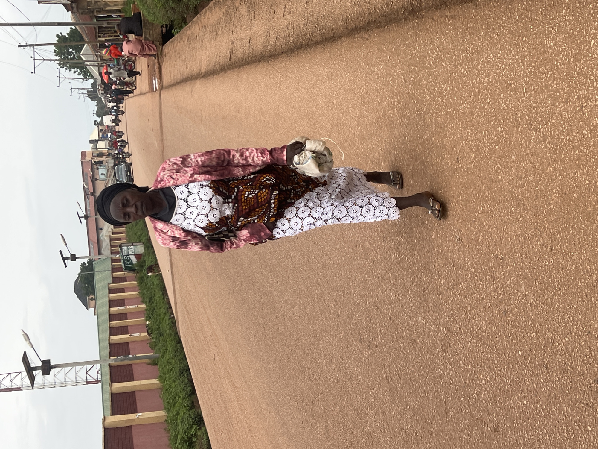 A woman in a patterned dress and headscarf walks on a sandy road in Durumi, Nigeria. She carries a white bag over her shoulder. In the background, a bustling street scene unfolds with motorcycles, cars, and people. Buildings line the road, and power lines stretch across the sky. The sky appears overcast, suggesting a cloudy day. The image captures a typical street scene in an urban or semi-urban area of Nigeria, with a focus on the woman as the central subject amidst the lively activity. The road is paved with a coarse, light brown material. The overall impression is one of everyday life and movement. There is no visible text in the image.