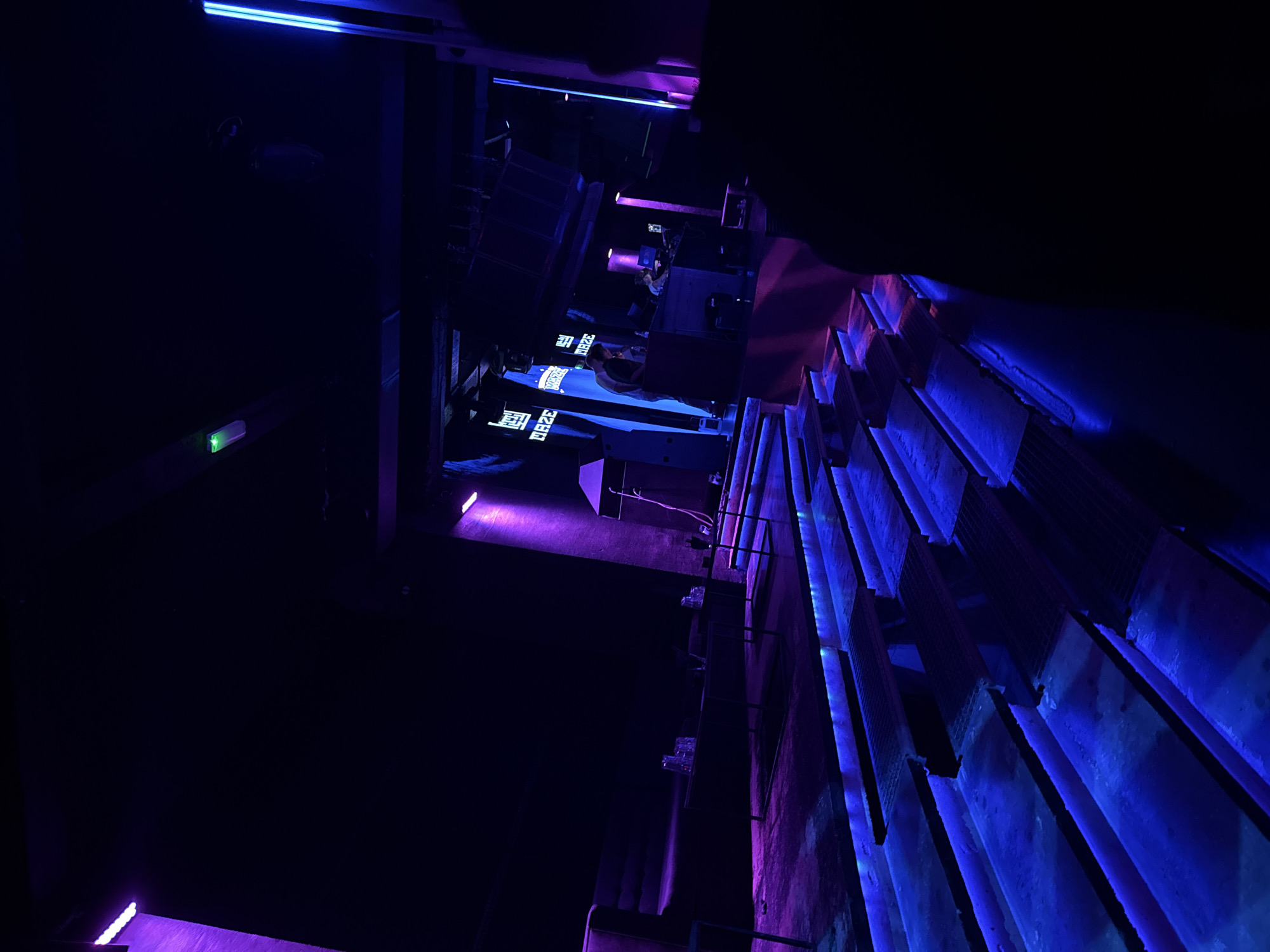 The image shows a low-angle, dimly lit interior in Varna, Bulgaria, featuring an industrial-themed setting with blue and purple lighting. A horizontal structure spans the upper section with illuminated text that is partially legible, possibly a sign or display. Below this, a shelf-like structure is mounted on a wall, holding several glasses and what may be bottles. To the right, stacked rectangular forms, potentially concrete or stone elements, cast long shadows under the blue lights. On the far left, near the top, is a security camera. In the background, there is a silhouette of a person on what looks like a platform. The image emphasizes textures and a dark, futuristic atmosphere.